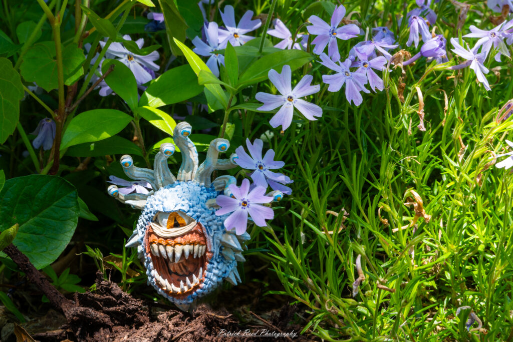 "A whimsical depiction of a beholder floating among a vibrant array of flowers. The beholder's large, central eye and multiple eyestalks are intricately detailed, contrasting with the colorful petals and lush greenery surrounding it. The scene captures a fantastical atmosphere, blending the menacing presence of the beholder with the delicate beauty of nature."