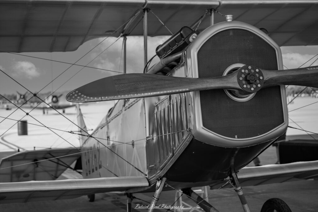 "A vintage bi-plane captured in striking black and white, showcasing its classic design and nostalgic charm. The aircraft features two stacked wings, a propeller at the front, and a distinctive rounded fuselage. The image highlights the intricate details of the bi-plane, including its wire supports and exposed framework, set against a dramatic sky or a scenic backdrop. The monochrome effect enhances the timeless quality of the aircraft, evoking a sense of early aviation history and adventure. This portrait captures the elegance and beauty of bi-plane design, emphasizing its place in aviation heritage."