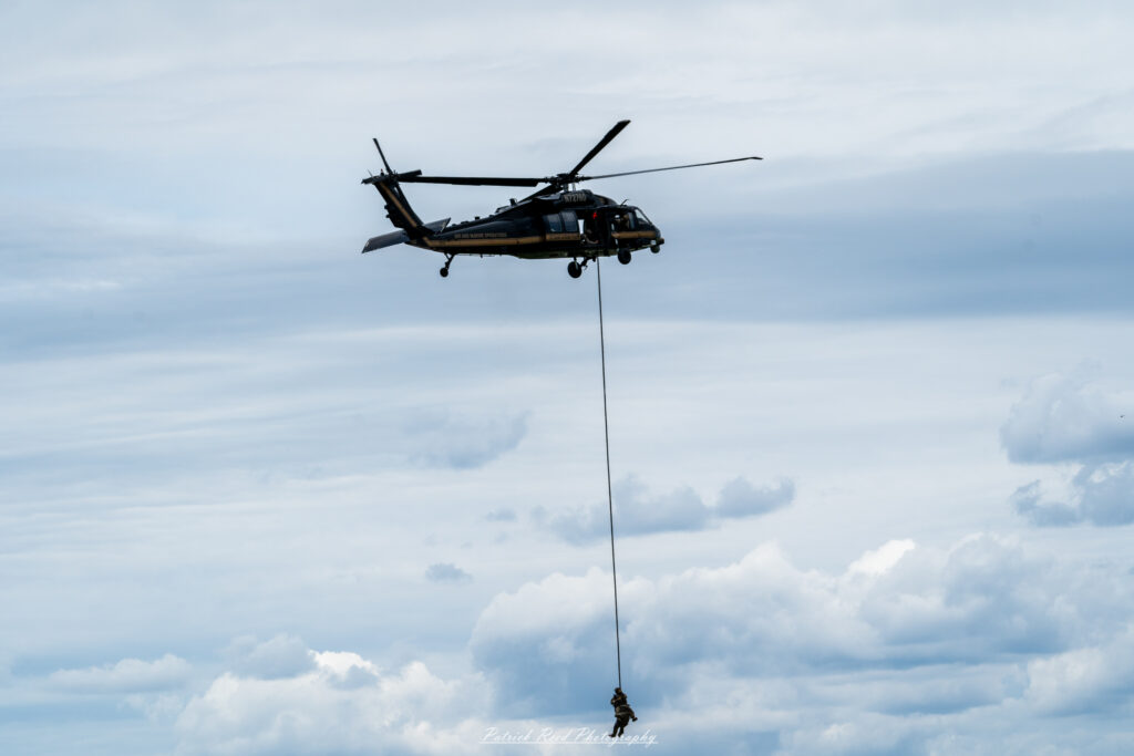 "A Black Hawk helicopter in flight, transporting two soldiers suspended below it via cable. The helicopter's sleek black design and powerful rotor blades create a dynamic scene against a clear blue sky. The soldiers, equipped with tactical gear and harnesses, are safely secured as they are lifted from the ground, showcasing the helicopter's capability for aerial extraction and transport. The backdrop may include a rugged terrain or battlefield setting, highlighting the versatility and strength of the Black Hawk in military operations and rescue missions."