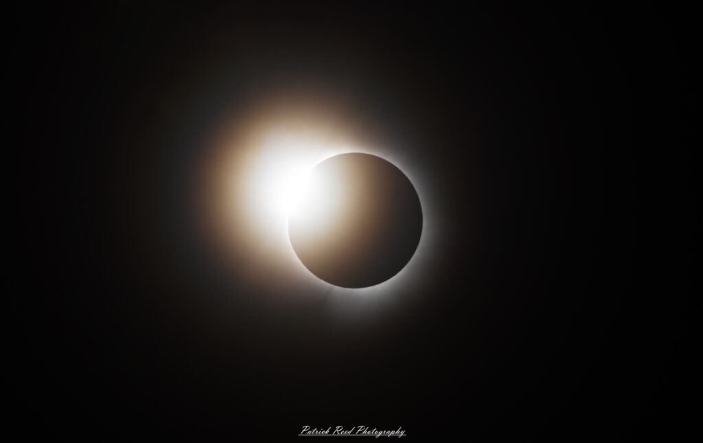 "The 'Diamond Ring' effect of a solar eclipse, a breathtaking moment just before and after totality. In this image, the last sliver of the sun is visible as a bright, glowing point of light, resembling a sparkling diamond on a ring formed by the darkened moon. Surrounding the 'diamond,' the solar corona begins to emerge, creating a stunning contrast between light and shadow. The sky around the eclipse is a deep, twilight blue, enhancing the dramatic visual impact of this rare astronomical phenomenon."