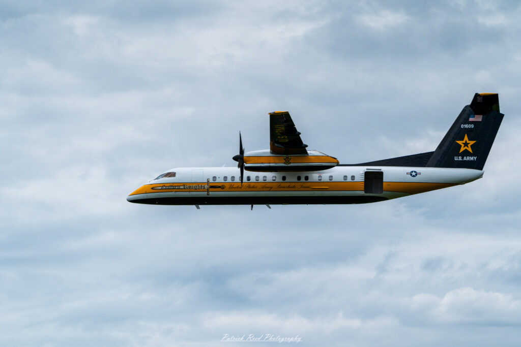"The U.S. Army Golden Knights transport plane, a sleek C-31 Troopship, shown in flight as it prepares to drop parachutists for an aerial demonstration. The aircraft features the distinctive Golden Knights logo prominently displayed on its fuselage, showcasing the team’s colors of black and gold. The plane's large cargo door is open, revealing parachutists poised to exit, ready for their precision jumps. Against a backdrop of clear blue skies, the scene captures the excitement and anticipation of the jump, highlighting the aircraft's role in facilitating thrilling skydiving performances by the elite Army parachute team."