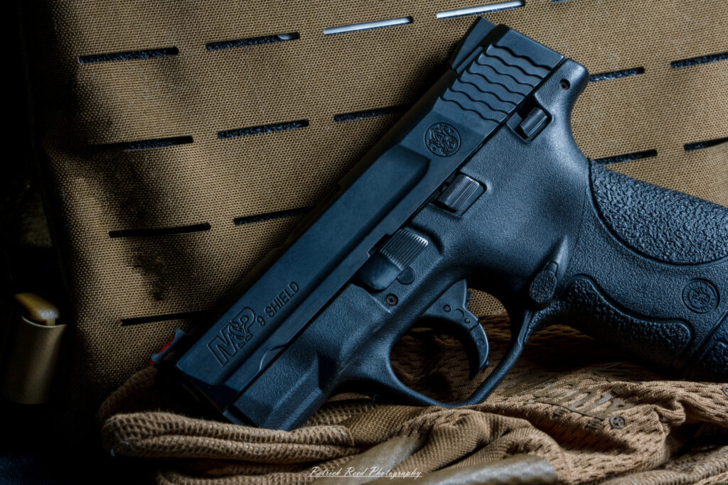 "A Smith & Wesson M&P Shield pistol leaning against a set of body armor, creating a scene that emphasizes readiness and protection. The contrast between the sleek design of the handgun and the rugged, tactical look of the body armor highlights the gear's purpose in personal defense and law enforcement."