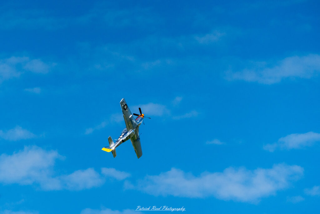 "The P-51D Mustang, an iconic World War II fighter aircraft, is showcased in a captivating profile view during flight. This vintage warplane features a sleek, elongated fuselage and a distinctive bubble canopy that provides excellent visibility for the pilot. The P-51D is painted in a vibrant color scheme, often displaying military markings, including invasion stripes and unit insignia. Known for its exceptional speed and maneuverability, the Mustang is powered by a powerful Rolls-Royce Merlin engine. The image captures the P-51D's historical significance and beauty, highlighting its role as a key fighter in achieving air superiority during the war."