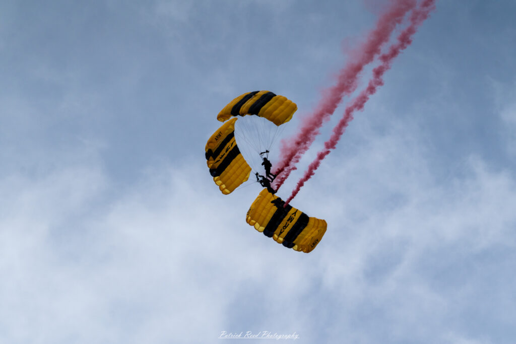 "Three parachutists in a dynamic formation during a skydive, captured mid-air against a bright blue sky. Each skydiver wears a brightly colored jumpsuit and helmet, showcasing their unique designs as they maintain precise positions in the formation. The parachutists are linked together by their arms and legs, displaying incredible teamwork and coordination. Below them, the landscape is visible, creating a stunning backdrop for this thrilling aerial activity. The scene highlights the exhilaration and camaraderie of skydiving, emphasizing the skill involved in maintaining a stable and synchronized formation during free fall."