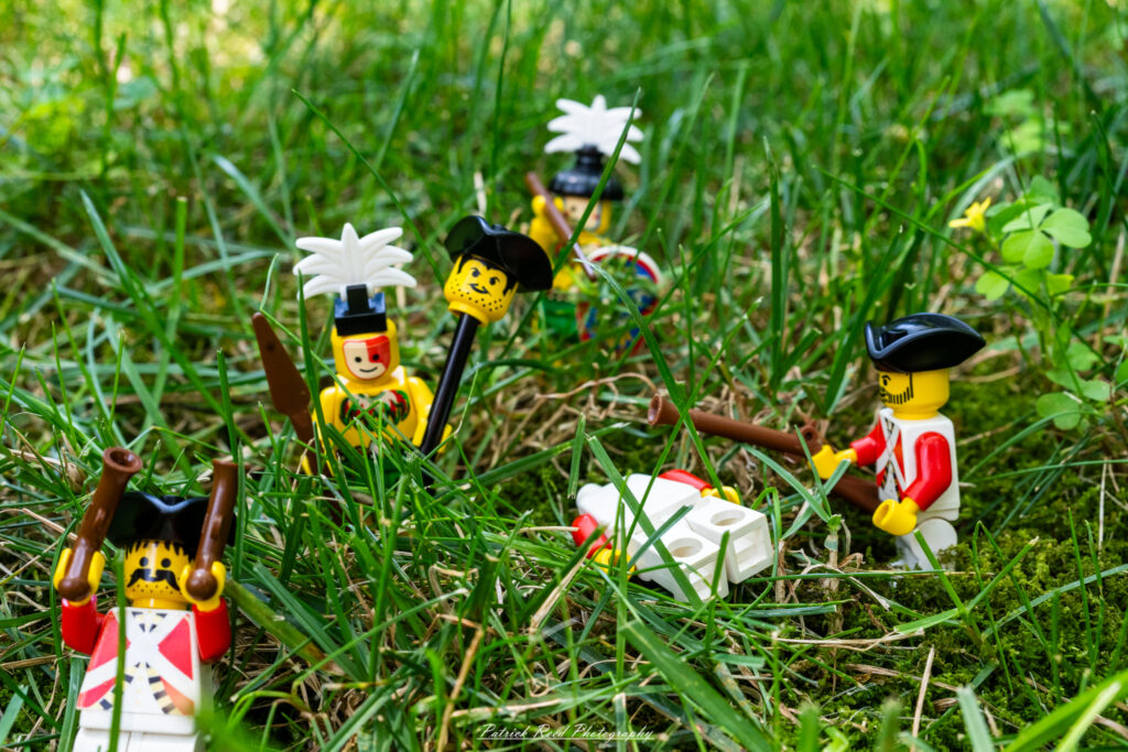 "LEGO figures of Native American warriors engaging in a dramatic scene where they are depicted as overcoming toy soldiers. The vibrant colors of the LEGO pieces create a playful yet intense atmosphere, with the warriors in dynamic poses, wielding traditional weapons. The toy soldiers, in their classic military attire, appear to be in the midst of a playful battle, set against a backdrop of a LEGO landscape that enhances the imaginative play."