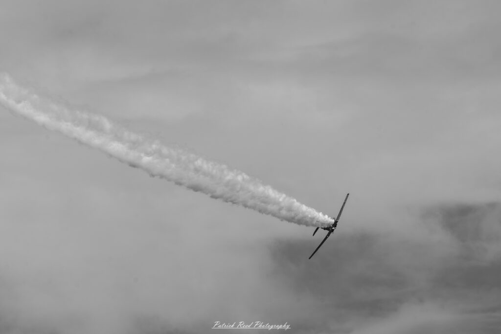 "A T-6 Texan, the classic single-engine advanced trainer aircraft from World War II, shown in flight. The plane features a low-wing monoplane design with a distinctive round engine cowling and a two-seat tandem cockpit. Its metallic body is often adorned with military insignia, and the plane’s tail and wingtips might feature bright yellow or red stripes, characteristic of training aircraft. The T-6 Texan is celebrated for its role in training thousands of pilots, and its presence evokes a sense of aviation history and the legacy of WWII."