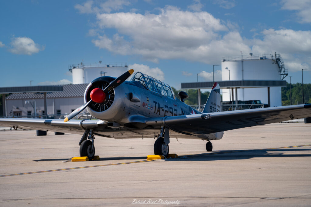"A T-6 Texan, a classic trainer aircraft, is positioned on the tarmac, showcasing its distinctive design and vibrant color scheme. The aircraft features a sleek, rounded fuselage with a prominent canopy and twin tail fins. Its polished wings and landing gear are detailed against the backdrop of a clear blue sky. Sunlight reflects off the aircraft's surface, highlighting its vintage charm and historical significance as a training plane for many military pilots. The tarmac is lined with service equipment and possibly other aircraft in the background, emphasizing the bustling atmosphere of an airfield."