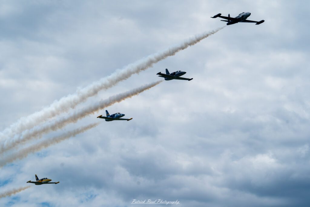 "Four vintage jets flying in tight formation against a clear blue sky, showcasing their classic designs and historical significance. Each aircraft, painted in vibrant colors and military markings from their respective eras, exhibits unique features, such as rounded noses, distinct tail designs, and sleek wings. The jets maintain precise spacing and altitude, creating a striking visual display that emphasizes teamwork and precision flying. This formation flight highlights the beauty and nostalgia of vintage military aviation, evoking memories of past conflicts and the evolution of aircraft technology."