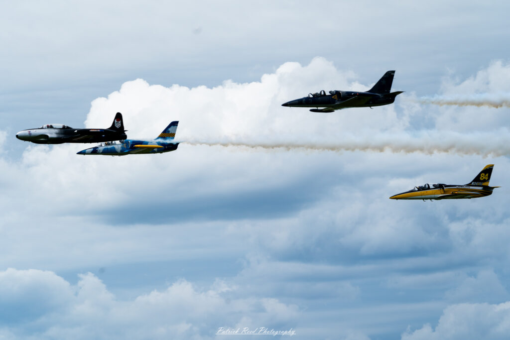 "Four vintage jets flying in tight formation against a clear blue sky, showcasing their classic designs and historical significance. Each aircraft, painted in vibrant colors and military markings from their respective eras, exhibits unique features, such as rounded noses, distinct tail designs, and sleek wings. The jets maintain precise spacing and altitude, creating a striking visual display that emphasizes teamwork and precision flying. This formation flight highlights the beauty and nostalgia of vintage military aviation, evoking memories of past conflicts and the evolution of aircraft technology."