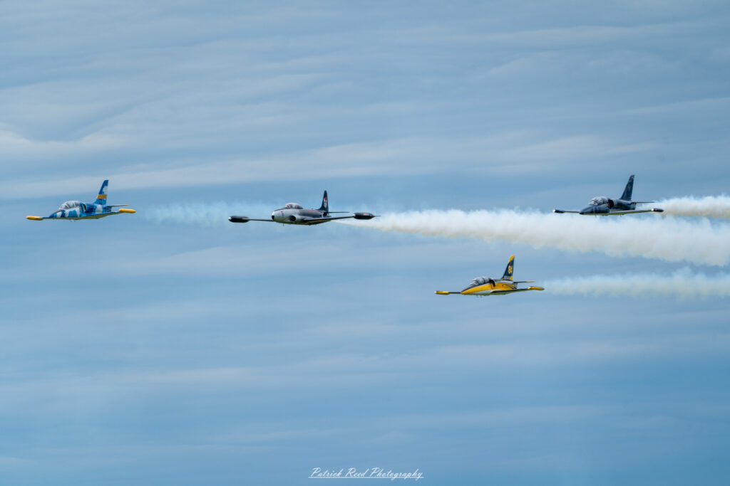 "Four vintage jets flying in tight formation against a clear blue sky, showcasing their classic designs and historical significance. Each aircraft, painted in vibrant colors and military markings from their respective eras, exhibits unique features, such as rounded noses, distinct tail designs, and sleek wings. The jets maintain precise spacing and altitude, creating a striking visual display that emphasizes teamwork and precision flying. This formation flight highlights the beauty and nostalgia of vintage military aviation, evoking memories of past conflicts and the evolution of aircraft technology."