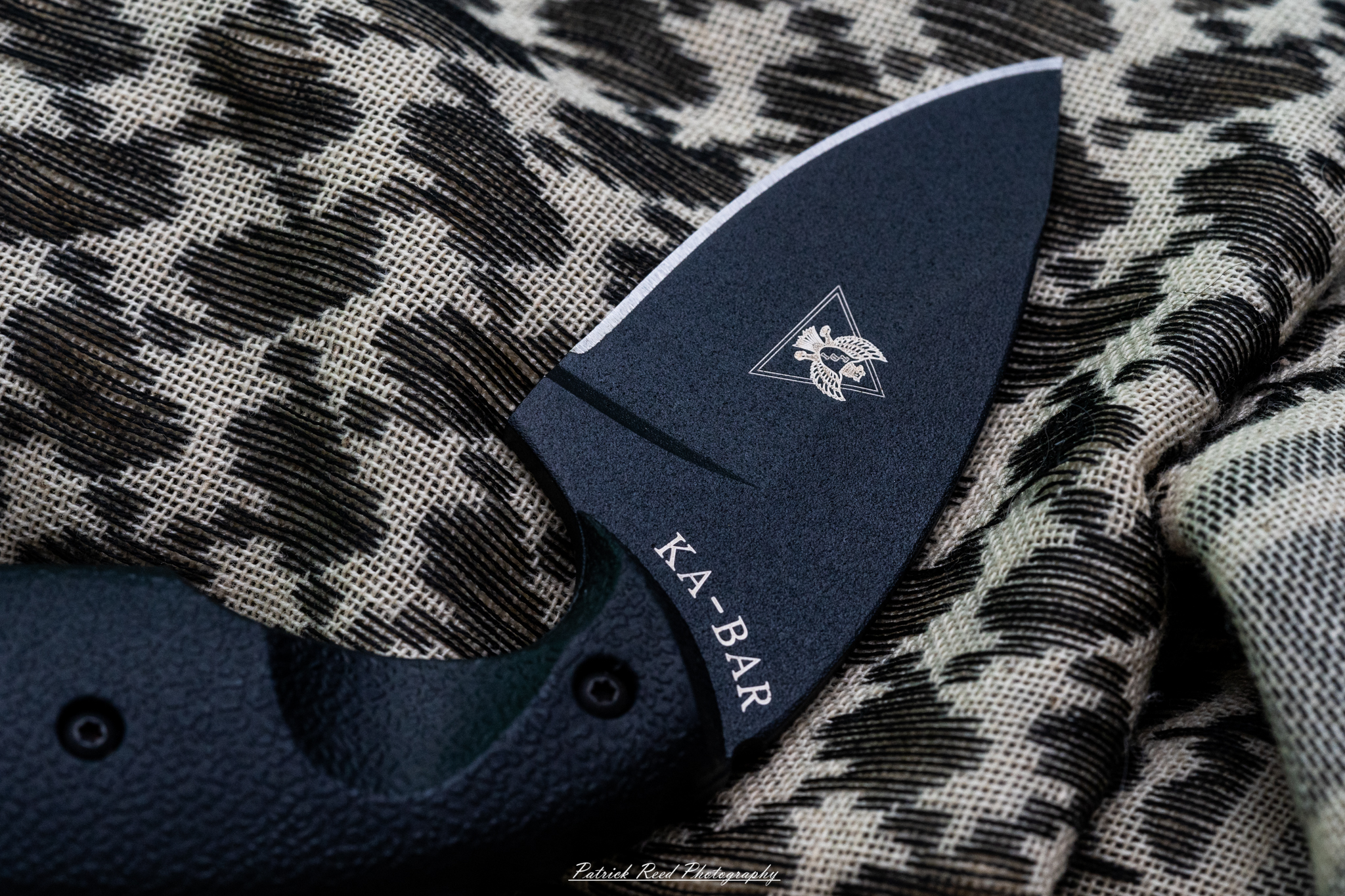 "A KA-BAR TDI knife resting on a wooden surface alongside a shemagh. The shemagh, with its intricate patterns and texture, adds an element of cultural significance to the scene. The knife's sharp blade and sturdy handle contrast with the softness of the fabric, emphasizing the practicality of both items in outdoor and tactical settings."