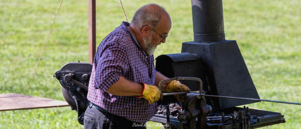 A blacksmith sawing metal in his workshop, focused on the task at hand. He uses a large hand saw, applying steady pressure as he cuts through a piece of metal. The scene showcases the skill and dedication of the blacksmith, with tools and materials surrounding him, reflecting the craftsmanship of traditional metalworking.