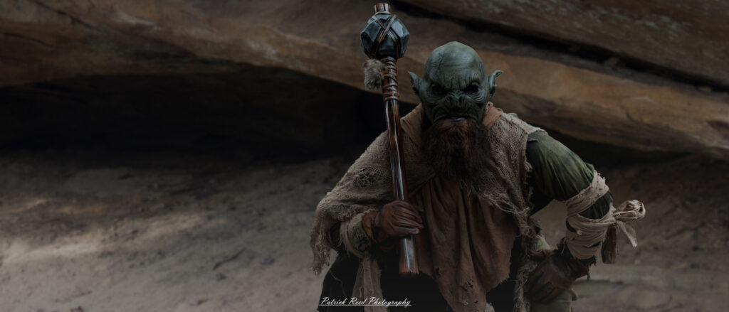 "A small, sneaky goblin stands at the entrance of a dark cave, its pointed ears twitching as it scans the surroundings. The goblin's sharp teeth and glowing eyes are illuminated by the dim light from the cave behind it, casting eerie shadows on its leathery skin. Its hunched posture and ragged clothing add to its mischievous and dangerous demeanor, while the cave mouth looms ominously, hinting at the dangers lurking within."