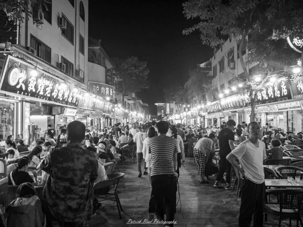"An open-air food market in China is bustling with activity, showcasing a vibrant array of fresh produce, spices, and street food. Vendors behind their stalls are busy preparing and selling various dishes, while customers browse and interact, creating a lively atmosphere. The market is filled with colorful fruits, vegetables, and aromatic ingredients, reflecting the rich culinary culture of the region. Overhead, signs in Chinese characters and bright awnings add to the visual appeal, while the scent of freshly cooked food wafts through the air. The scene is a snapshot of daily life, highlighting the community and tradition centered around food in China."