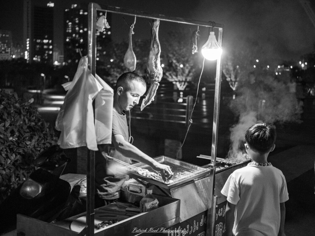Black-and-white photograph of a street vendor preparing skewers at a lamp-lit grill cart while a child stands beside rising smoke.