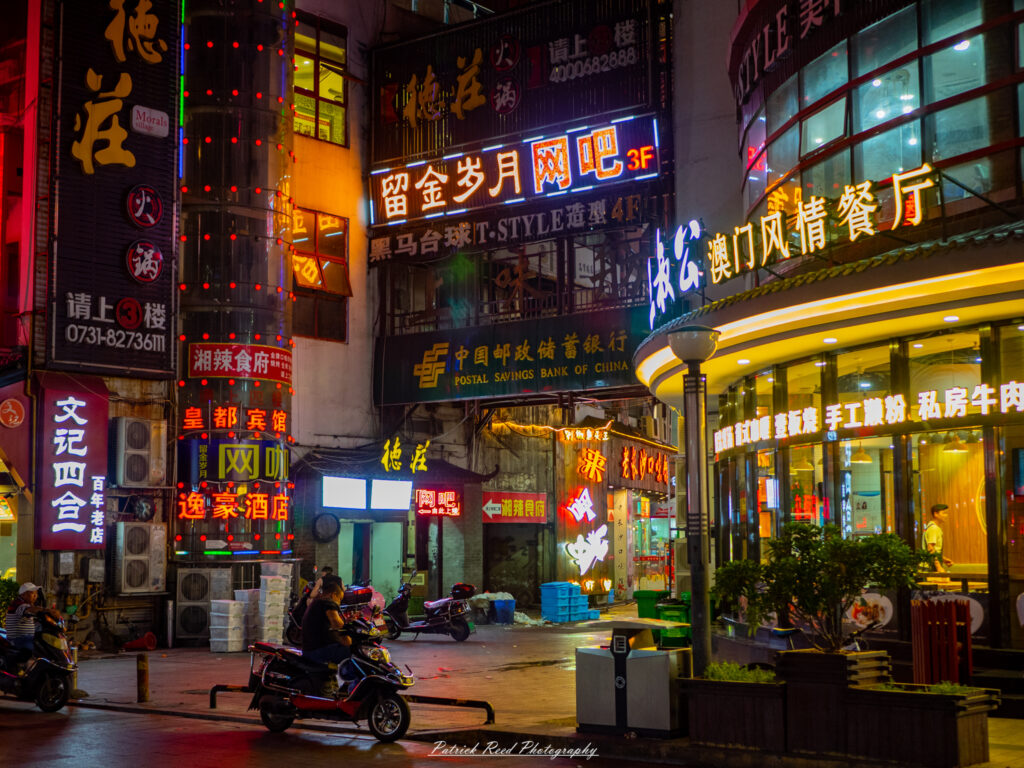 "A vibrant black and white image captures the bustling atmosphere of a Chinese street adorned with glowing neon signs. The signs, displaying a mix of Chinese characters and colorful graphics, illuminate the night, creating a dynamic contrast against the dark urban backdrop. Street vendors and pedestrians navigate through the lively scene, with the neon lights reflecting off nearby surfaces. This photograph encapsulates the energy and vibrancy of urban life in China, showcasing the cultural fusion of tradition and modernity."