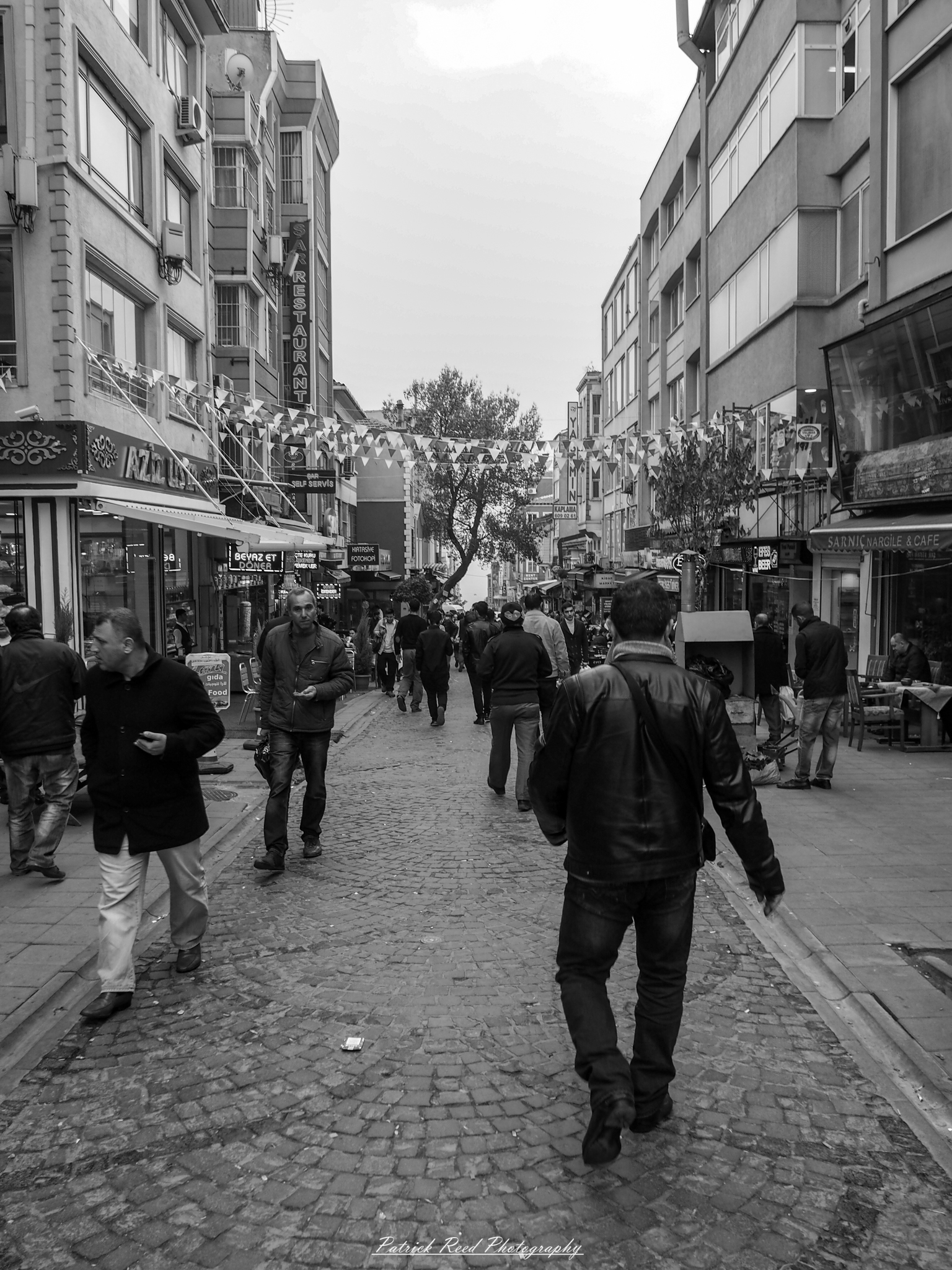 Istanbul Street Scene "A black and white image of a busy street in Istanbul, capturing the dynamic energy of the city. The scene features a mix of locals and tourists navigating the bustling thoroughfare, surrounded by historic architecture and shops. The absence of color emphasizes the contrasts between light and shadow, adding a timeless quality to the image. People engage in conversation, and the atmosphere is filled with a sense of movement and life, showcasing Istanbul's rich cultural tapestry in monochrome."