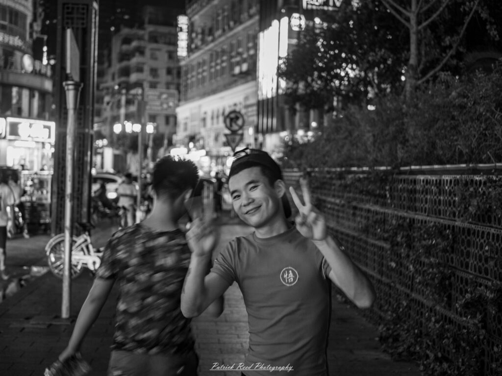 "A young Chinese man flashes a peace sign with a cheerful smile in a black and white photograph. The contrast of the monochrome tones emphasizes the warmth of his expression and the playful gesture. His casual attire and relaxed posture capture a sense of youth and joy. The background is softly blurred, directing the focus to his lively face and friendly demeanor. This image embodies a spirit of positivity and camaraderie, showcasing a moment of lightheartedness in everyday life."