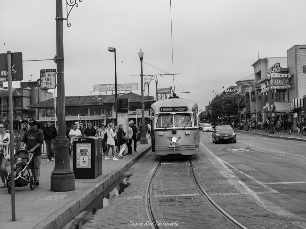 "A black and white image features a classic San Francisco trolley parked on the street, exuding a sense of nostalgia and timeless charm. The iconic trolley, with its distinct design and vintage details, stands still against a backdrop of historic buildings and urban scenery. The monochrome tones enhance the textures of the trolley and the cobblestone street, while shadows play across the scene, adding depth and character. This stationary trolley captures the essence of San Francisco’s rich history and serves as a striking visual reminder of the city’s iconic transportation."