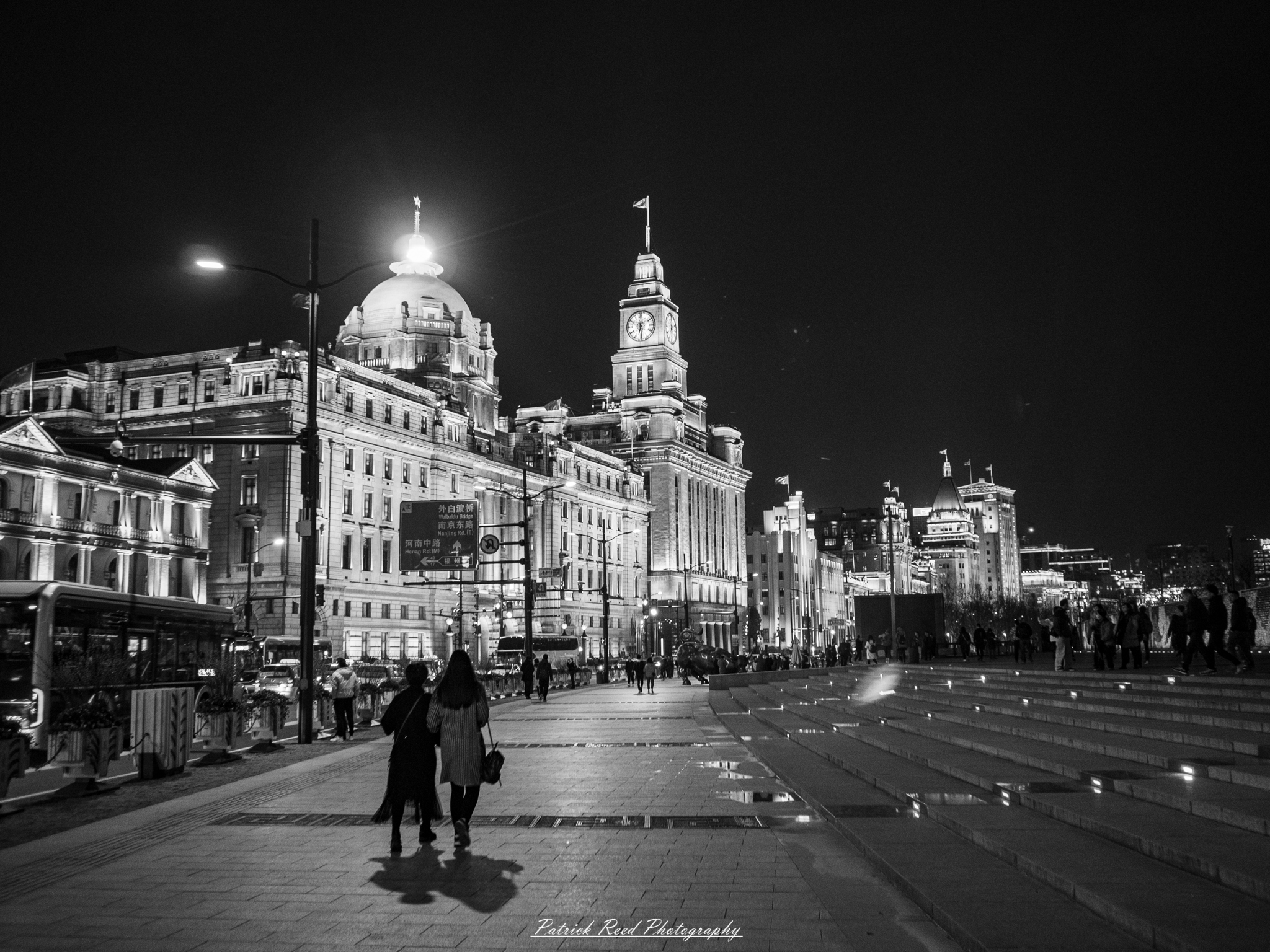The Bund "A striking black and white scene featuring two women strolling along the Bund in Shanghai, China. The iconic skyline serves as a dramatic backdrop, showcasing the city's blend of historic architecture and modern skyscrapers. The women, dressed in stylish attire, walk closely together, perhaps deep in conversation, while the reflective pavement adds a sense of depth to the image. This timeless capture evokes a feeling of elegance and urban charm, highlighting the cultural richness of one of China's most famous waterfronts."