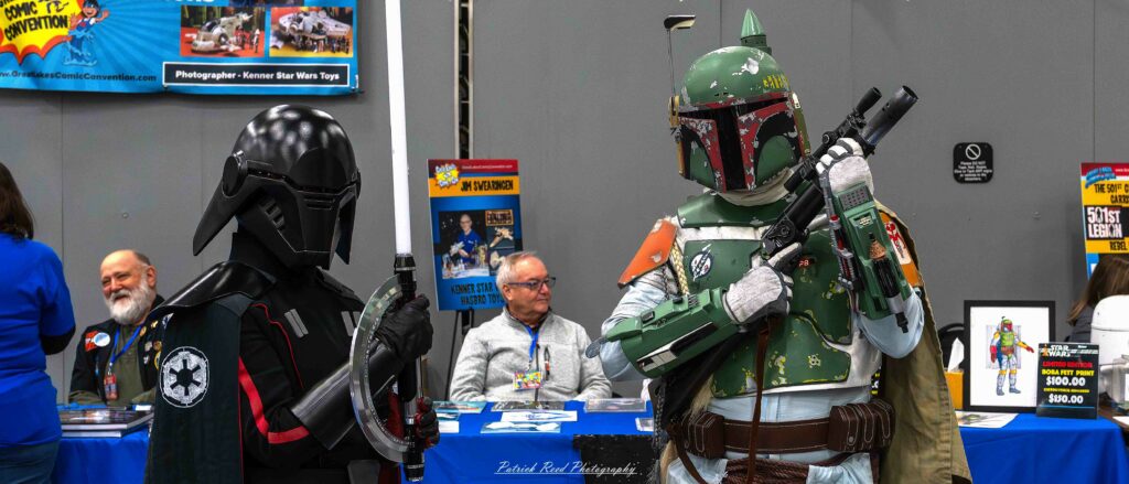 Two cosplayers dressed as Boba Fett and an Inquisitor from Star Wars pose together at the Great Lakes Comic Con cosplay contest. Boba Fett wears his iconic green Mandalorian armor with a dented helmet, equipped with blaster and jetpack. Next to him, the Inquisitor is clad in black robes, a red lightsaber in hand, and sporting a menacing helmet with angular features.