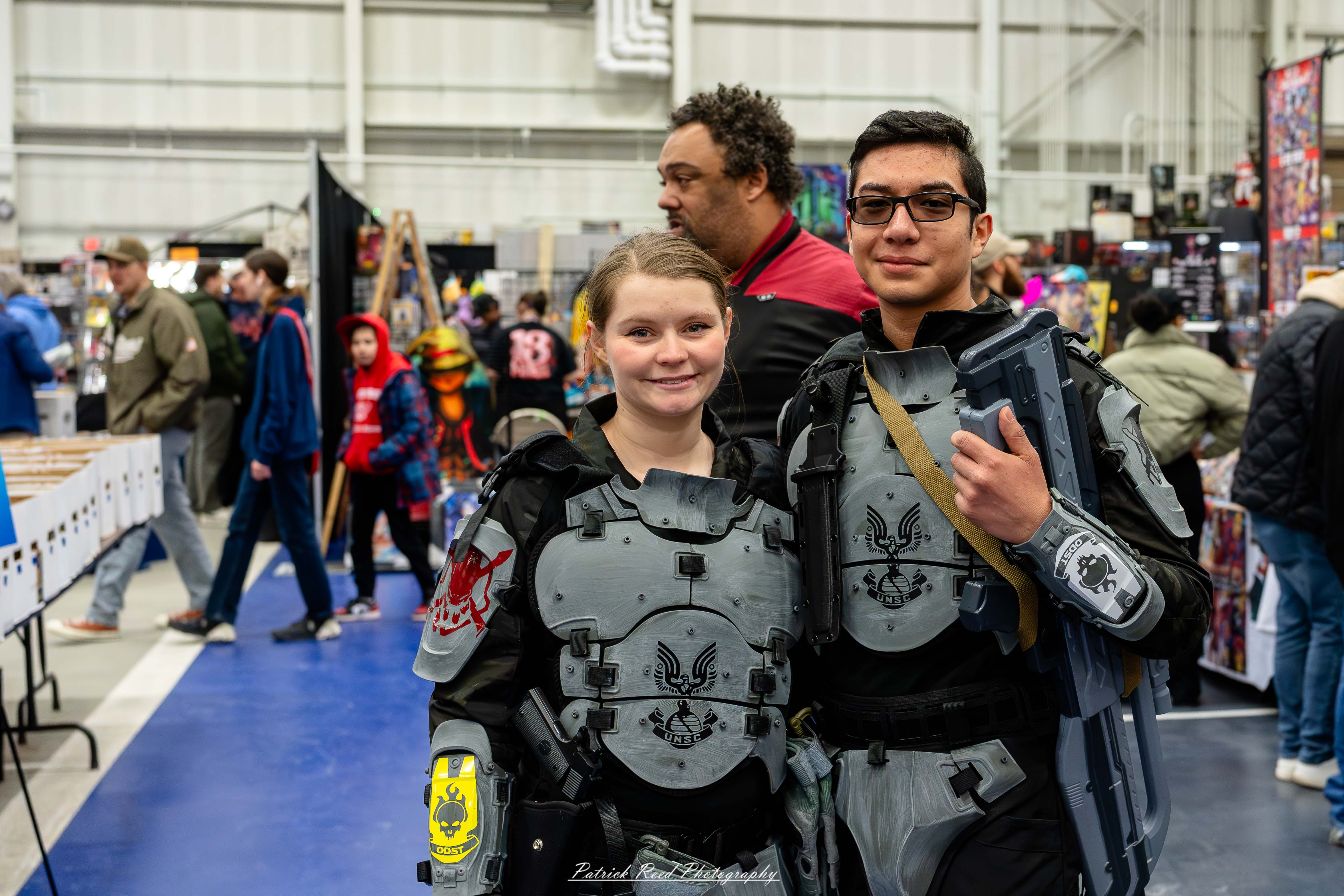 Great Lakes Comic Con 10 Alt text: A man and woman dressed in detailed Halo UNSC Trooper armor pose together at the Great Lakes Comic Con cosplay contest. Their futuristic combat suits feature green and gray plating with visored helmets, tactical gear, and replica weapons, capturing the look of soldiers from the Halo universe. Bright stage lights highlight the intricate armor as the crowd watches.