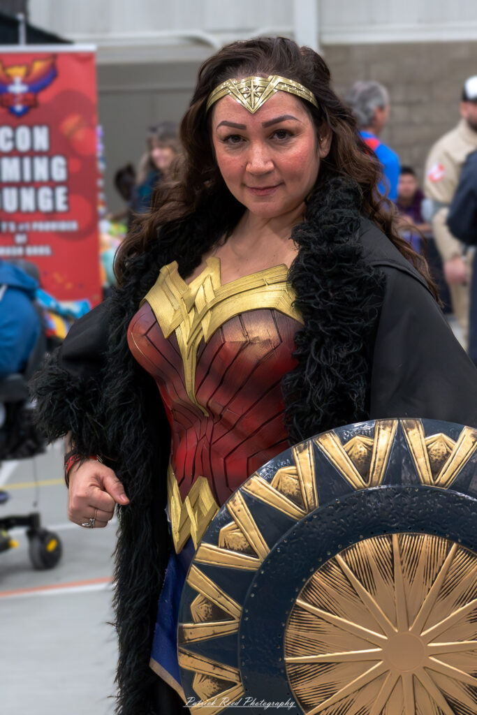 A cosplayer dressed as Wonder Woman stands confidently at the Great Lakes Comic Con cosplay contest. She wears Wonder Woman’s iconic red and gold armor, blue skirt with white stars, and wields a shield and lasso. Bright stage lights highlight the detailed craftsmanship as the audience looks on.