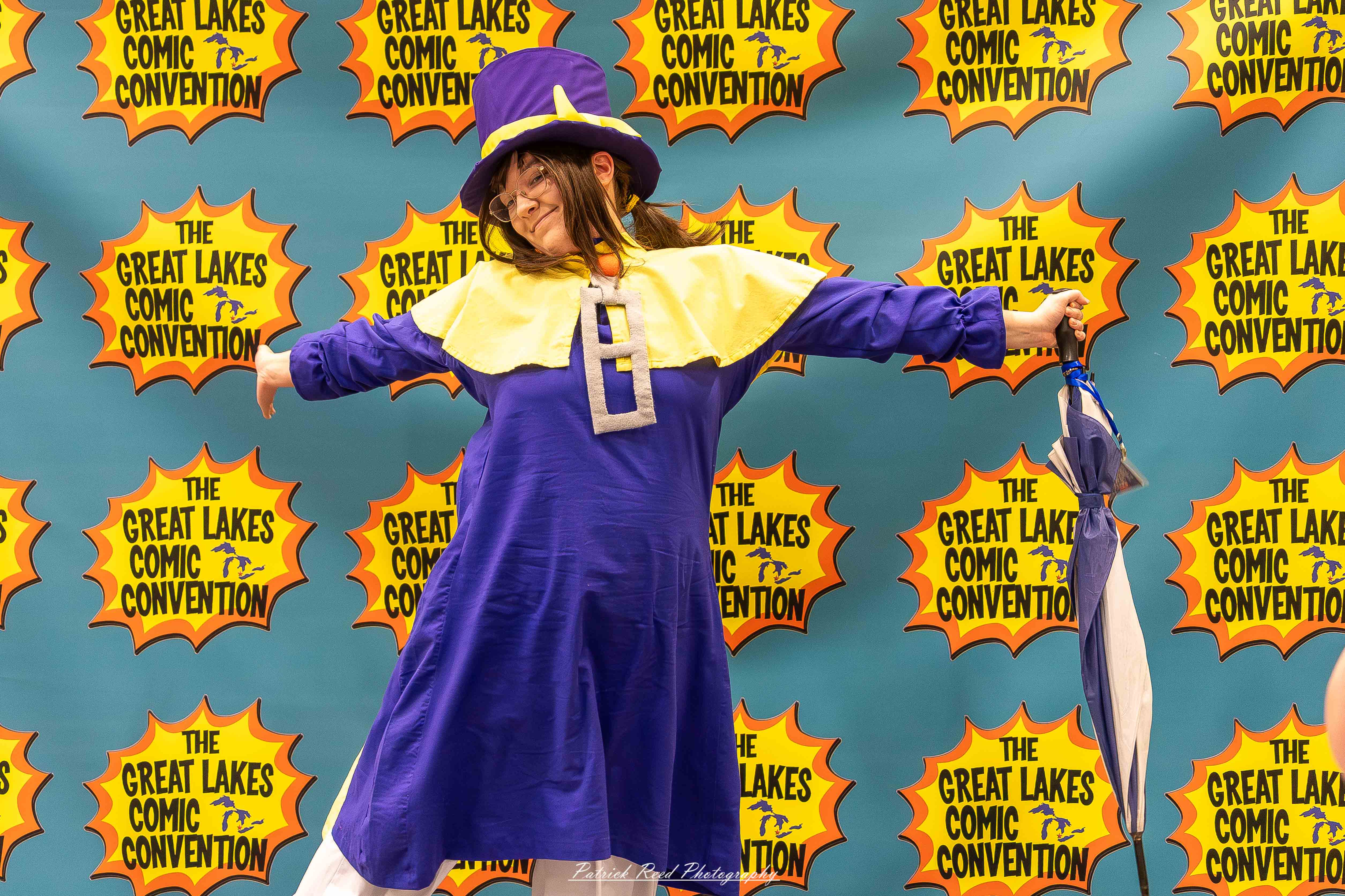 Great Lakes Comic Con 19 Alt text: A group of cosplayers posing on stage at the Great Lakes Comic Con cosplay contest, dressed as various characters from comics, anime, and video games. The audience watches from below as the participants show off their detailed costumes under bright stage lights.