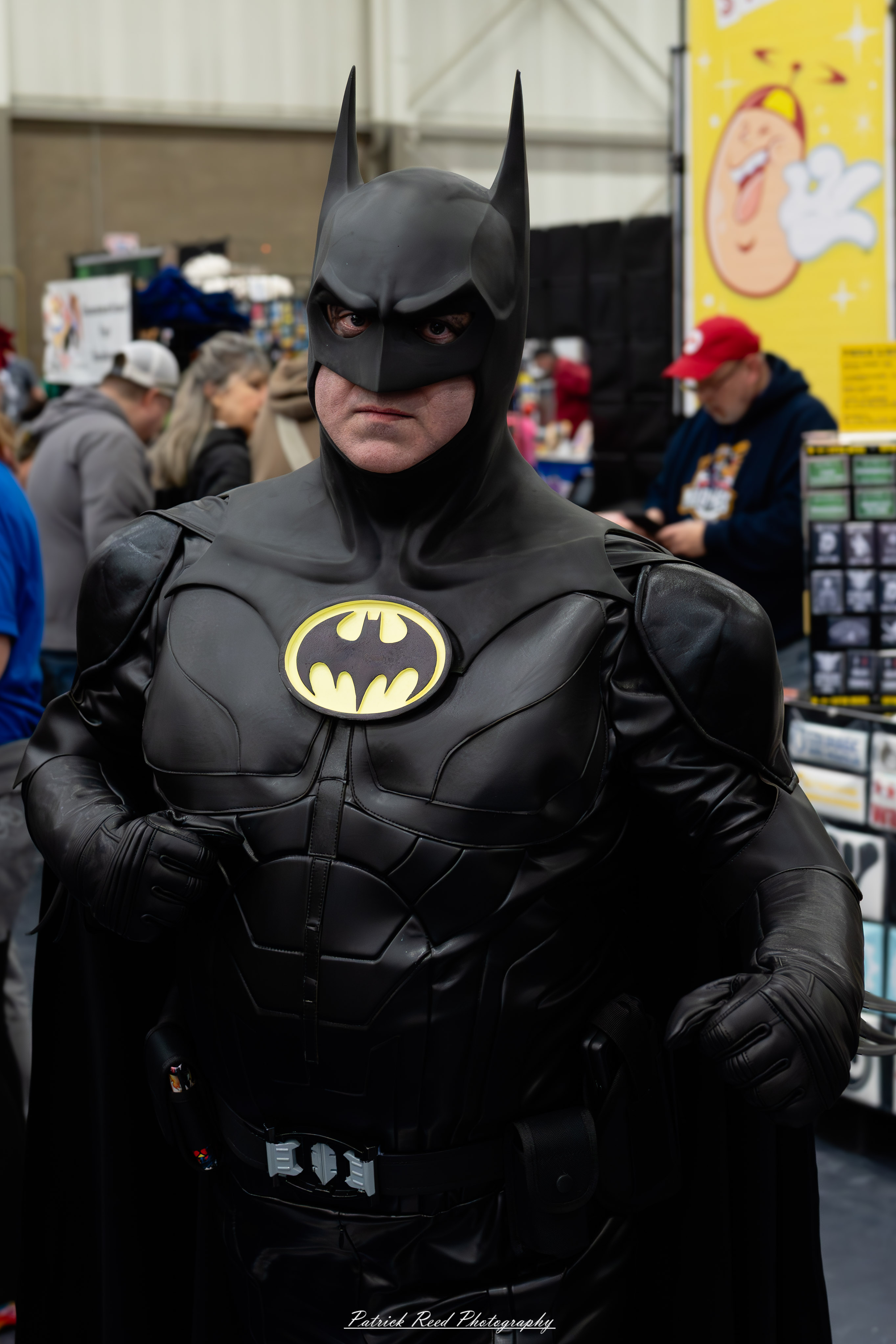 Great Lakes Comic Con 2 A cosplayer dressed as Batman stands confidently at the Great Lakes Comic Con cosplay contest. The costume features a dark gray bodysuit with a black bat emblem on the chest, a flowing cape, and a utility belt.