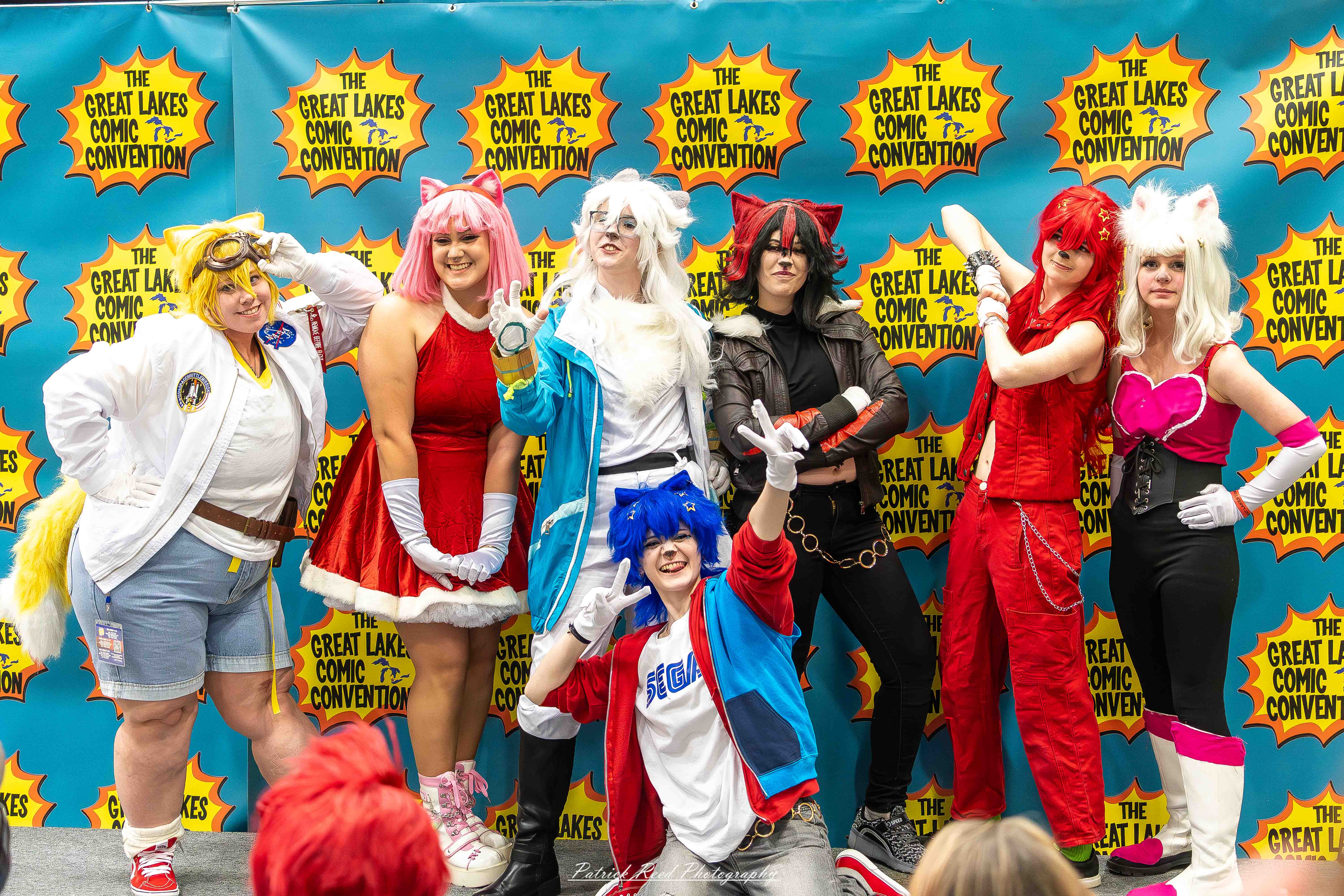 Great Lakes Comic Con 27 Alt text: A group of cosplayers posing on stage at the Great Lakes Comic Con cosplay contest, dressed as various characters from comics, anime, and video games. The audience watches from below as the participants show off their detailed costumes under bright stage lights.