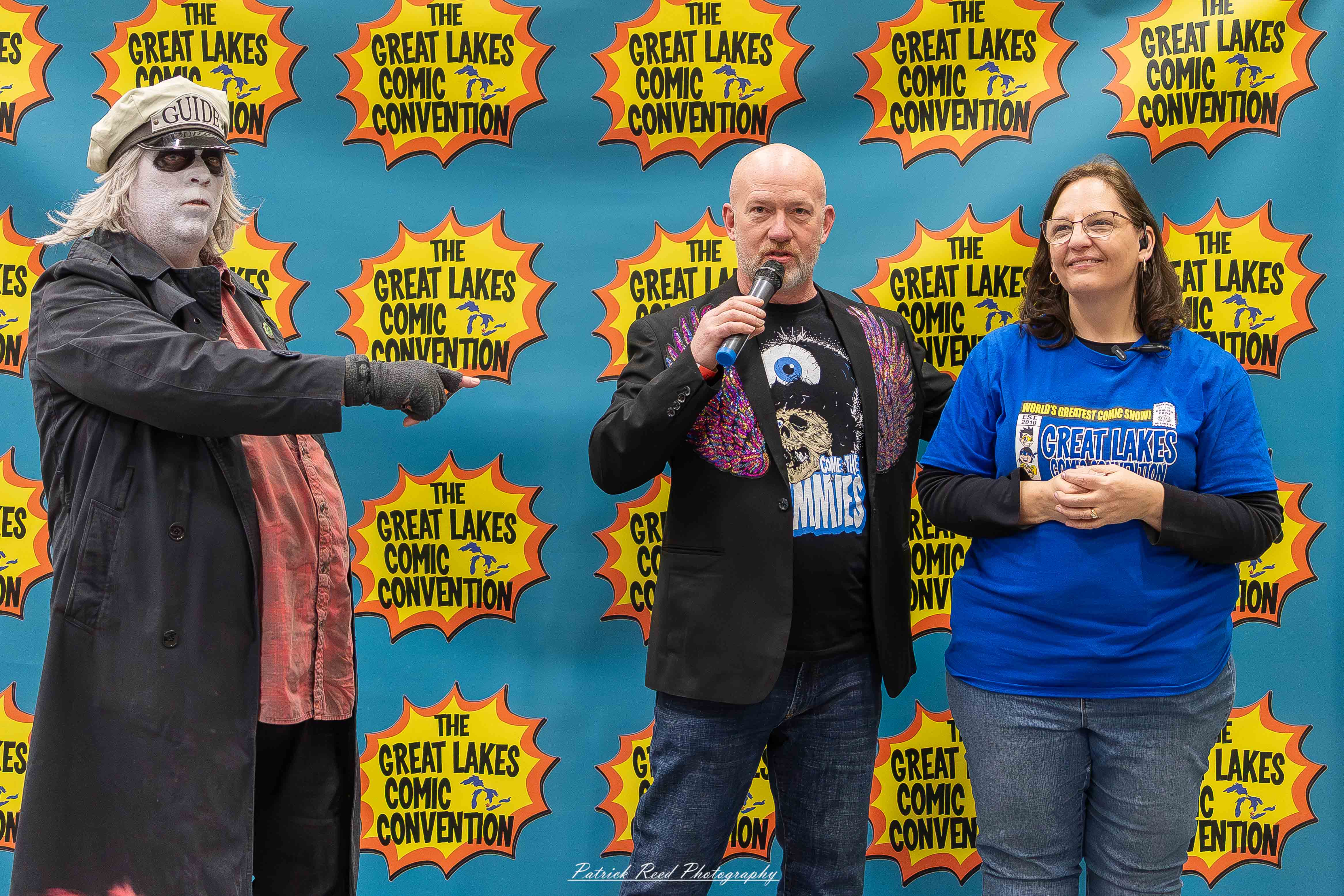Great Lakes Comic Con 47 Alt text: A group of cosplayers posing on stage at the Great Lakes Comic Con cosplay contest, dressed as various characters from comics, anime, and video games. The audience watches from below as the participants show off their detailed costumes under bright stage lights.