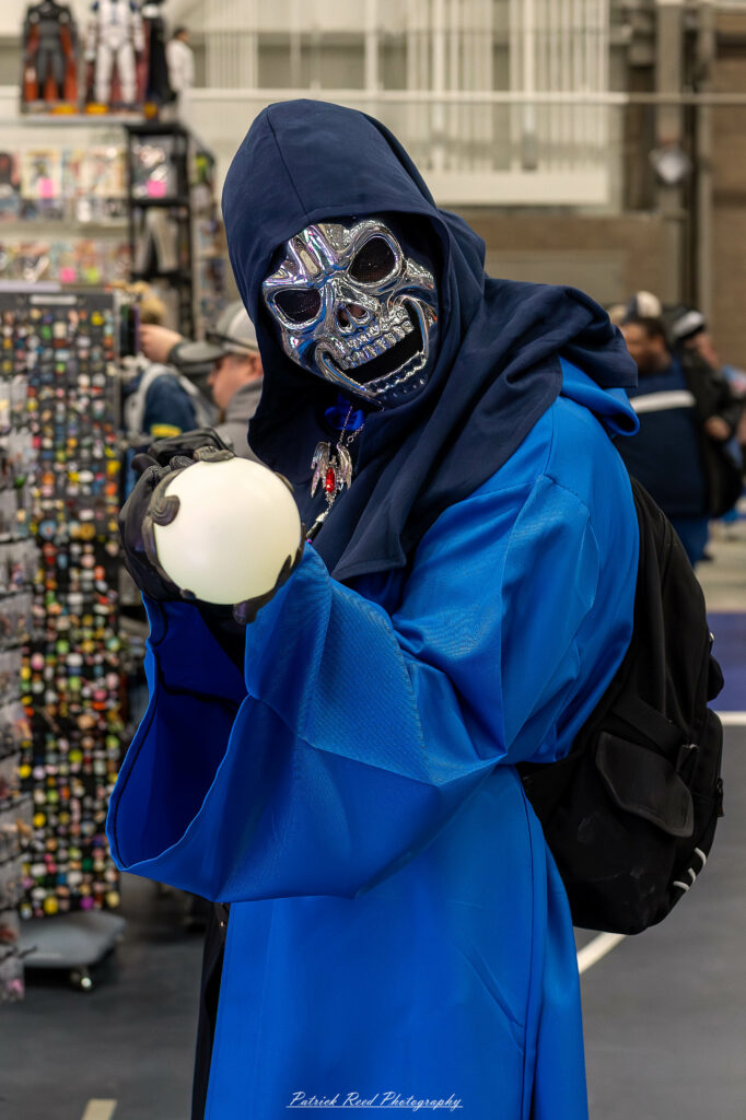 A cosplayer dressed as Skeletor from Masters of the Universe poses at the Great Lakes Comic Con cosplay contest. They wear a blue bodysuit with purple armor, a hooded cowl, and a skull mask. The cosplayer holds Skeletor’s iconic Havoc Staff topped with a ram’s skull,