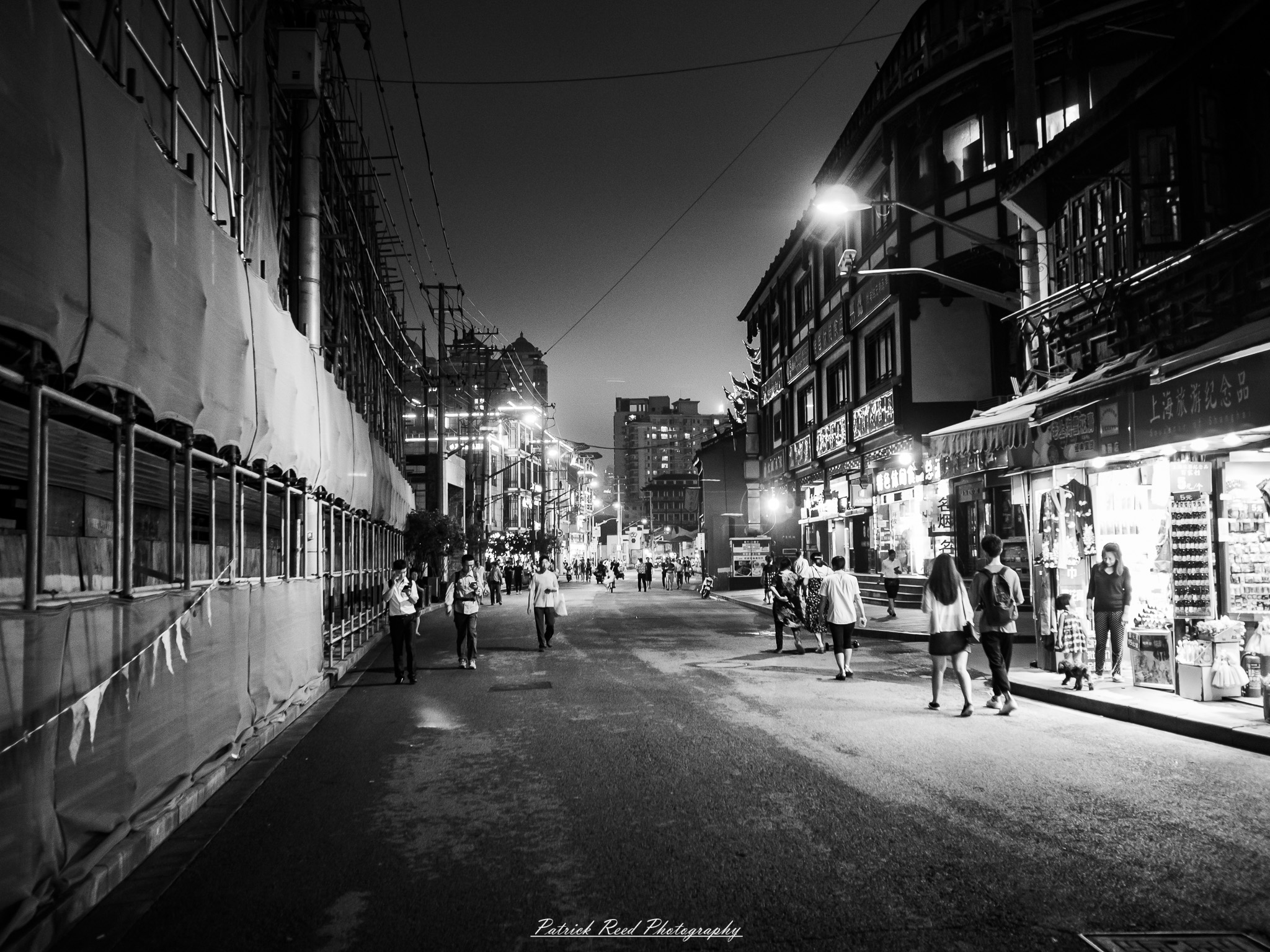 China Noir Scene 2 A noir-style street scene in China, featuring dimly lit, rain-soaked streets with reflections of neon signs. Traditional Chinese architecture blends with modern urban elements, including bright storefronts and illuminated signs. Silhouettes of people carrying umbrellas move through the misty atmosphere, creating a sense of mystery and cinematic drama. The wet pavement and dim lighting enhance the gritty, atmospheric vibe, reminiscent of classic film noir.