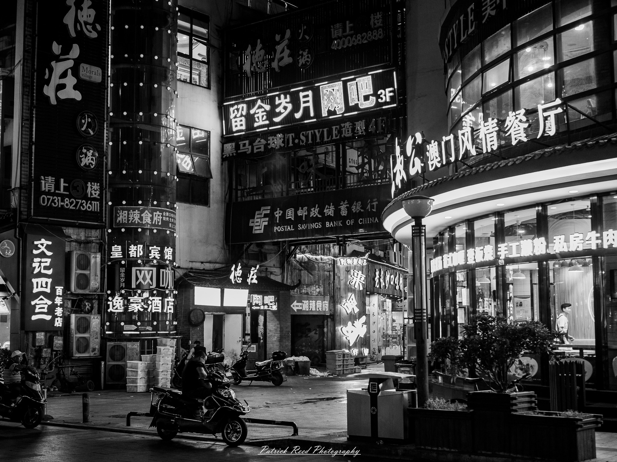 China Noir Scene 4 A noir-style scene of a narrow alley in China at night, with wet cobblestone pavement reflecting neon lights from signs and windows. A scooter is parked against a wall, partially illuminated by the glow of a flickering sign. The alley is framed by traditional Chinese architectural details, such as tiled roofs and lanterns, blending with urban elements like graffiti and wires. Silhouettes of people holding umbrellas walk through the misty atmosphere in the background, adding a sense of mystery and storytelling. The overall mood is cinematic, gritty, and evocative.