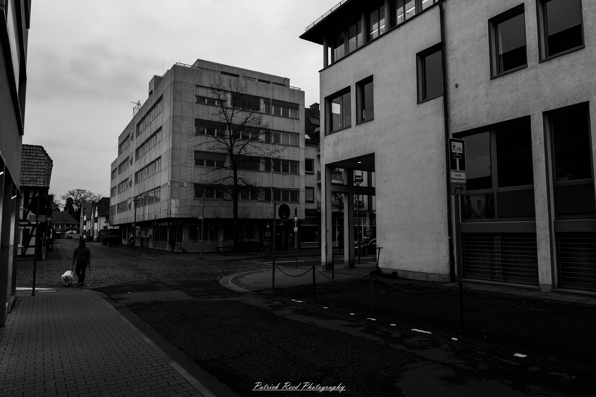 Russelsheim 1 A series of noir-style street photography scenes set in Rüsselsheim, Germany, at night. The dimly lit streets feature wet cobblestones reflecting the glow of streetlights and neon signs. Shadows and silhouettes of people, some walking or riding bicycles, add a mysterious atmosphere. Industrial architecture, such as factory buildings and modern urban structures, contrasts with old-world charm. Elements like parked cars, lampposts, and occasional graffiti create a gritty, cinematic ambiance. The misty air and moody lighting emphasize the noir aesthetic, evoking a sense of intrigue and storytelling.
