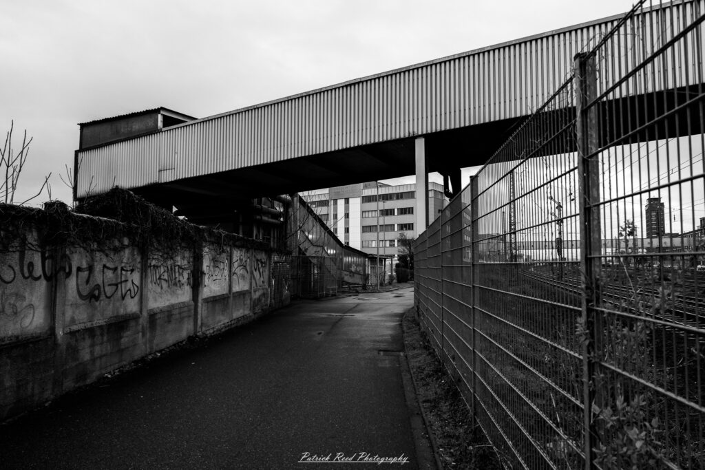 A series of noir-style street photography scenes set in Rüsselsheim, Germany, at night. The dimly lit streets feature wet cobblestones reflecting the glow of streetlights and neon signs. Shadows and silhouettes of people, some walking or riding bicycles, add a mysterious atmosphere. Industrial architecture, such as factory buildings and modern urban structures, contrasts with old-world charm. Elements like parked cars, lampposts, and occasional graffiti create a gritty, cinematic ambiance. The misty air and moody lighting emphasize the noir aesthetic, evoking a sense of intrigue and storytelling.
