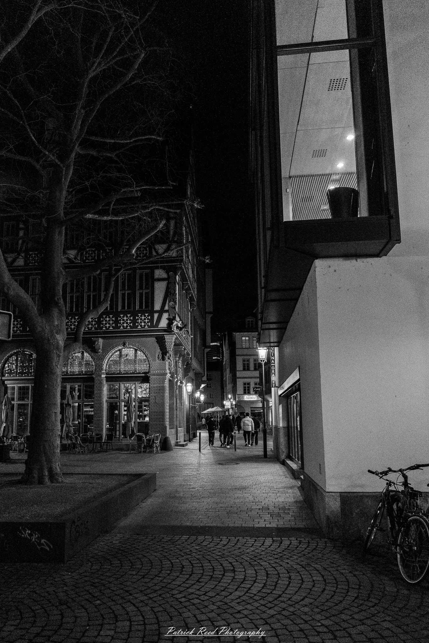 Russelsheim 11 A series of noir-style street photography scenes set in Rüsselsheim, Germany, at night. The dimly lit streets feature wet cobblestones reflecting the glow of streetlights and neon signs. Shadows and silhouettes of people, some walking or riding bicycles, add a mysterious atmosphere. Industrial architecture, such as factory buildings and modern urban structures, contrasts with old-world charm. Elements like parked cars, lampposts, and occasional graffiti create a gritty, cinematic ambiance. The misty air and moody lighting emphasize the noir aesthetic, evoking a sense of intrigue and storytelling.