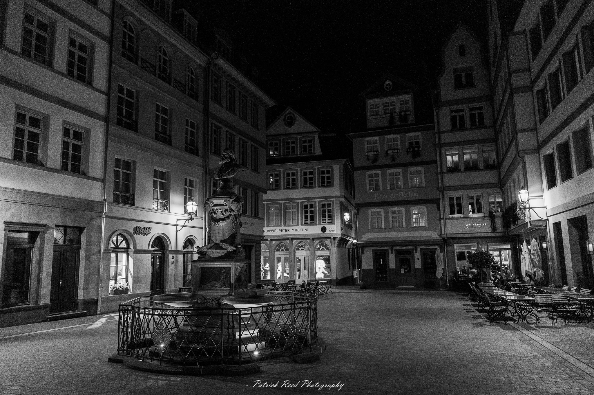 A series of noir-style street photography scenes set in Rüsselsheim, Germany, at night. The dimly lit streets feature wet cobblestones reflecting the glow of streetlights and neon signs. Shadows and silhouettes of people, some walking or riding bicycles, add a mysterious atmosphere. Industrial architecture, such as factory buildings and modern urban structures, contrasts with old-world charm. Elements like parked cars, lampposts, and occasional graffiti create a gritty, cinematic ambiance. The misty air and moody lighting emphasize the noir aesthetic, evoking a sense of intrigue and storytelling.