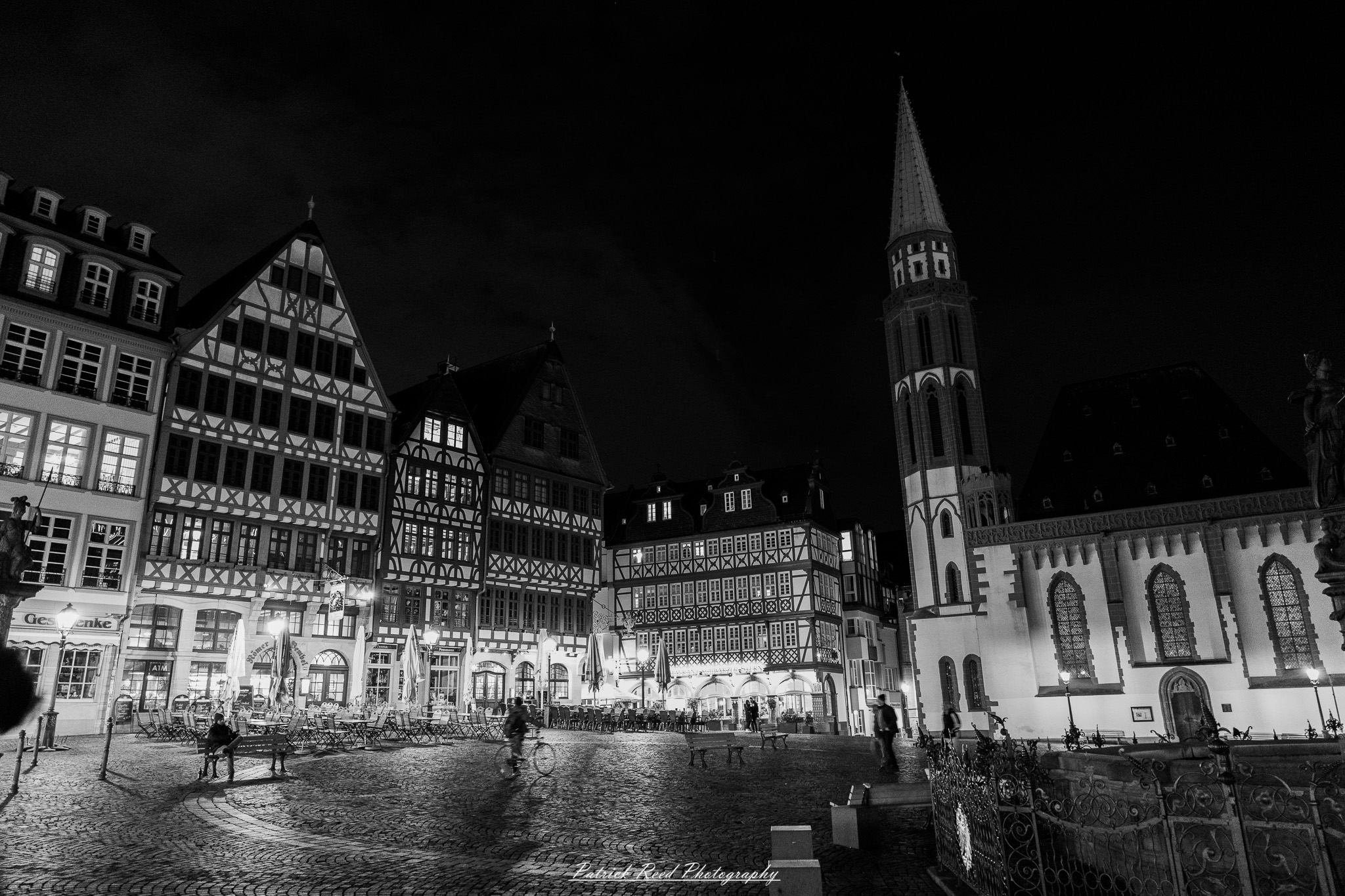 Russelsheim 14 A series of noir-style street photography scenes set in Rüsselsheim, Germany, at night. The dimly lit streets feature wet cobblestones reflecting the glow of streetlights and neon signs. Shadows and silhouettes of people, some walking or riding bicycles, add a mysterious atmosphere. Industrial architecture, such as factory buildings and modern urban structures, contrasts with old-world charm. Elements like parked cars, lampposts, and occasional graffiti create a gritty, cinematic ambiance. The misty air and moody lighting emphasize the noir aesthetic, evoking a sense of intrigue and storytelling.