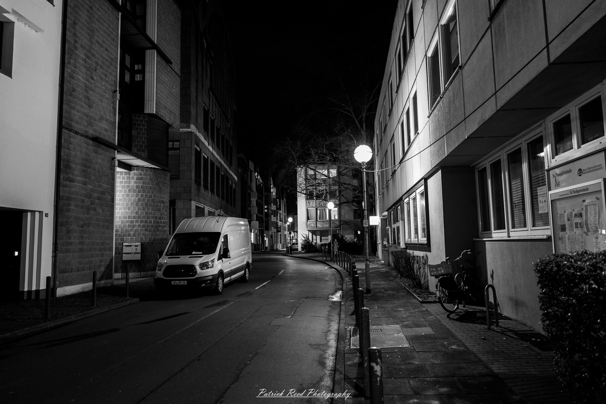 Russelsheim 16 A series of noir-style street photography scenes set in Rüsselsheim, Germany, at night. The dimly lit streets feature wet cobblestones reflecting the glow of streetlights and neon signs. Shadows and silhouettes of people, some walking or riding bicycles, add a mysterious atmosphere. Industrial architecture, such as factory buildings and modern urban structures, contrasts with old-world charm. Elements like parked cars, lampposts, and occasional graffiti create a gritty, cinematic ambiance. The misty air and moody lighting emphasize the noir aesthetic, evoking a sense of intrigue and storytelling.