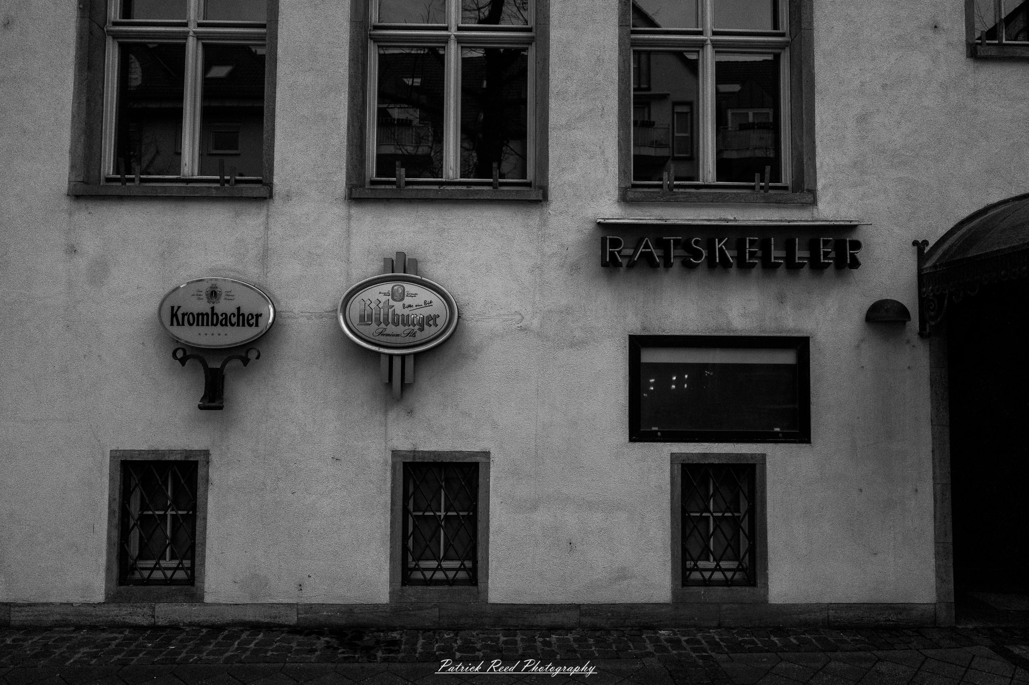 Russelsheim 18 A series of noir-style street photography scenes set in Rüsselsheim, Germany, at night. The dimly lit streets feature wet cobblestones reflecting the glow of streetlights and neon signs. Shadows and silhouettes of people, some walking or riding bicycles, add a mysterious atmosphere. Industrial architecture, such as factory buildings and modern urban structures, contrasts with old-world charm. Elements like parked cars, lampposts, and occasional graffiti create a gritty, cinematic ambiance. The misty air and moody lighting emphasize the noir aesthetic, evoking a sense of intrigue and storytelling.