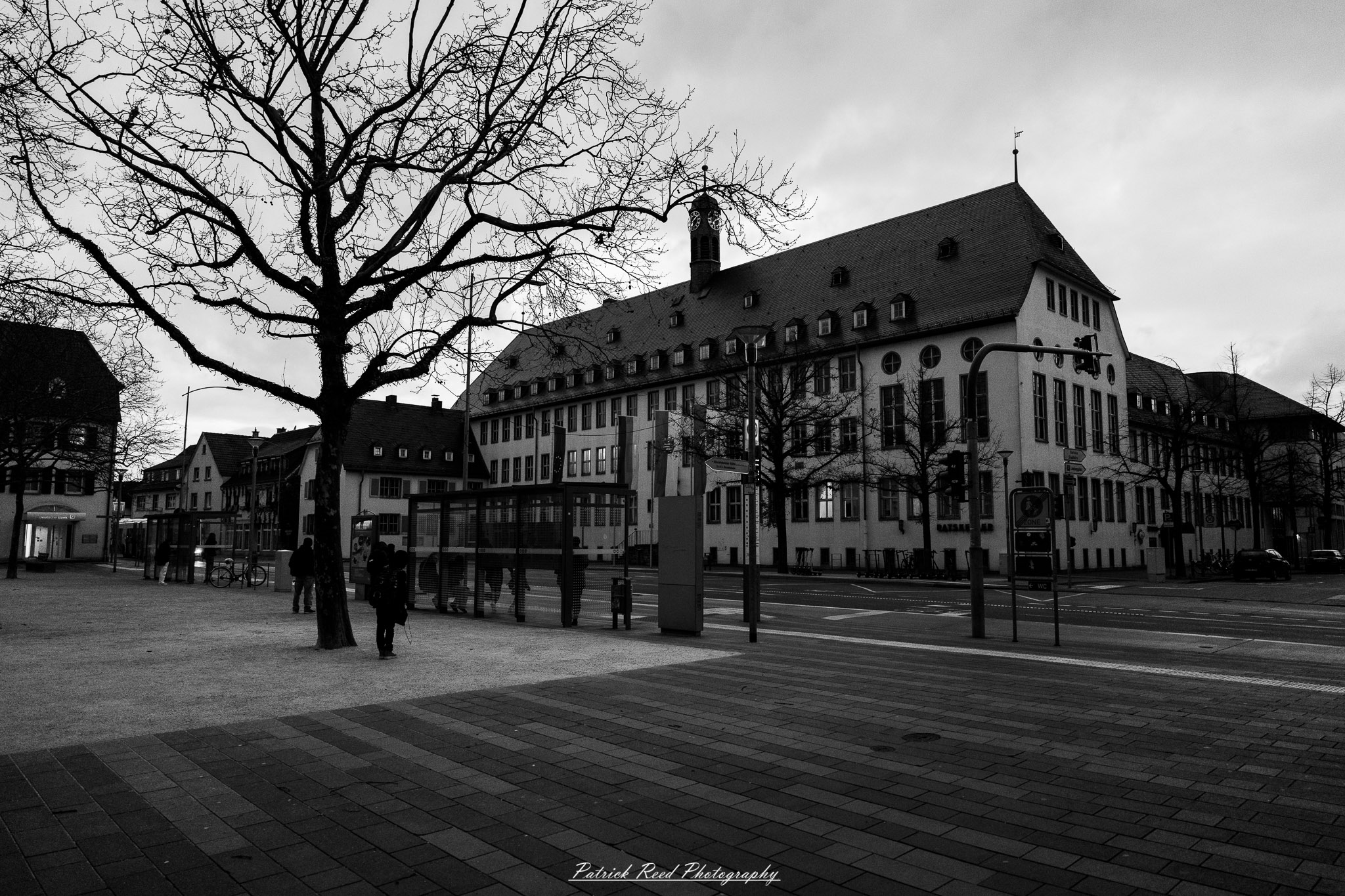 Russelsheim 19 A series of noir-style street photography scenes set in Rüsselsheim, Germany, at night. The dimly lit streets feature wet cobblestones reflecting the glow of streetlights and neon signs. Shadows and silhouettes of people, some walking or riding bicycles, add a mysterious atmosphere. Industrial architecture, such as factory buildings and modern urban structures, contrasts with old-world charm. Elements like parked cars, lampposts, and occasional graffiti create a gritty, cinematic ambiance. The misty air and moody lighting emphasize the noir aesthetic, evoking a sense of intrigue and storytelling.