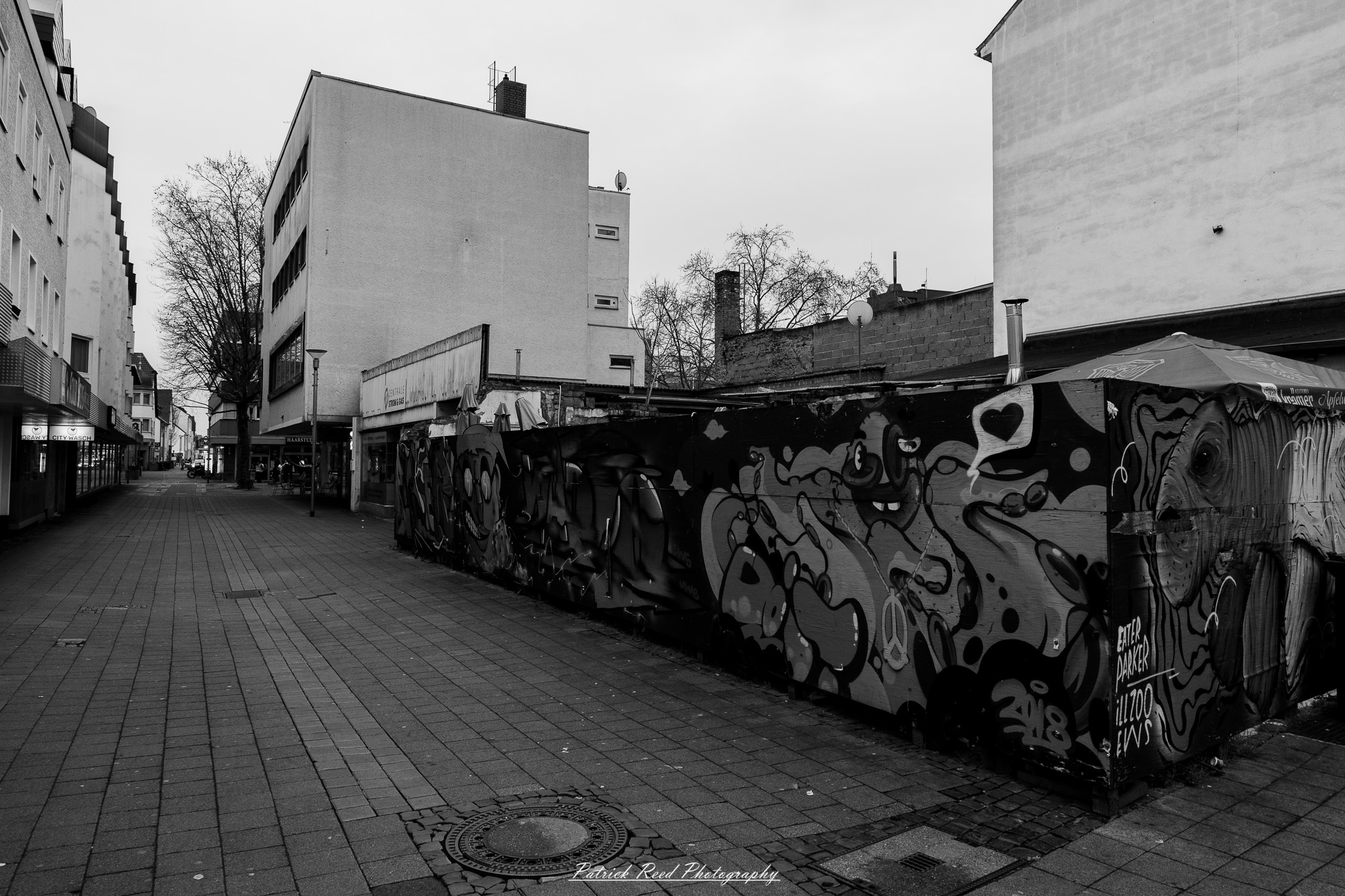 Russelsheim 2 A series of noir-style street photography scenes set in Rüsselsheim, Germany, at night. The dimly lit streets feature wet cobblestones reflecting the glow of streetlights and neon signs. Shadows and silhouettes of people, some walking or riding bicycles, add a mysterious atmosphere. Industrial architecture, such as factory buildings and modern urban structures, contrasts with old-world charm. Elements like parked cars, lampposts, and occasional graffiti create a gritty, cinematic ambiance. The misty air and moody lighting emphasize the noir aesthetic, evoking a sense of intrigue and storytelling.