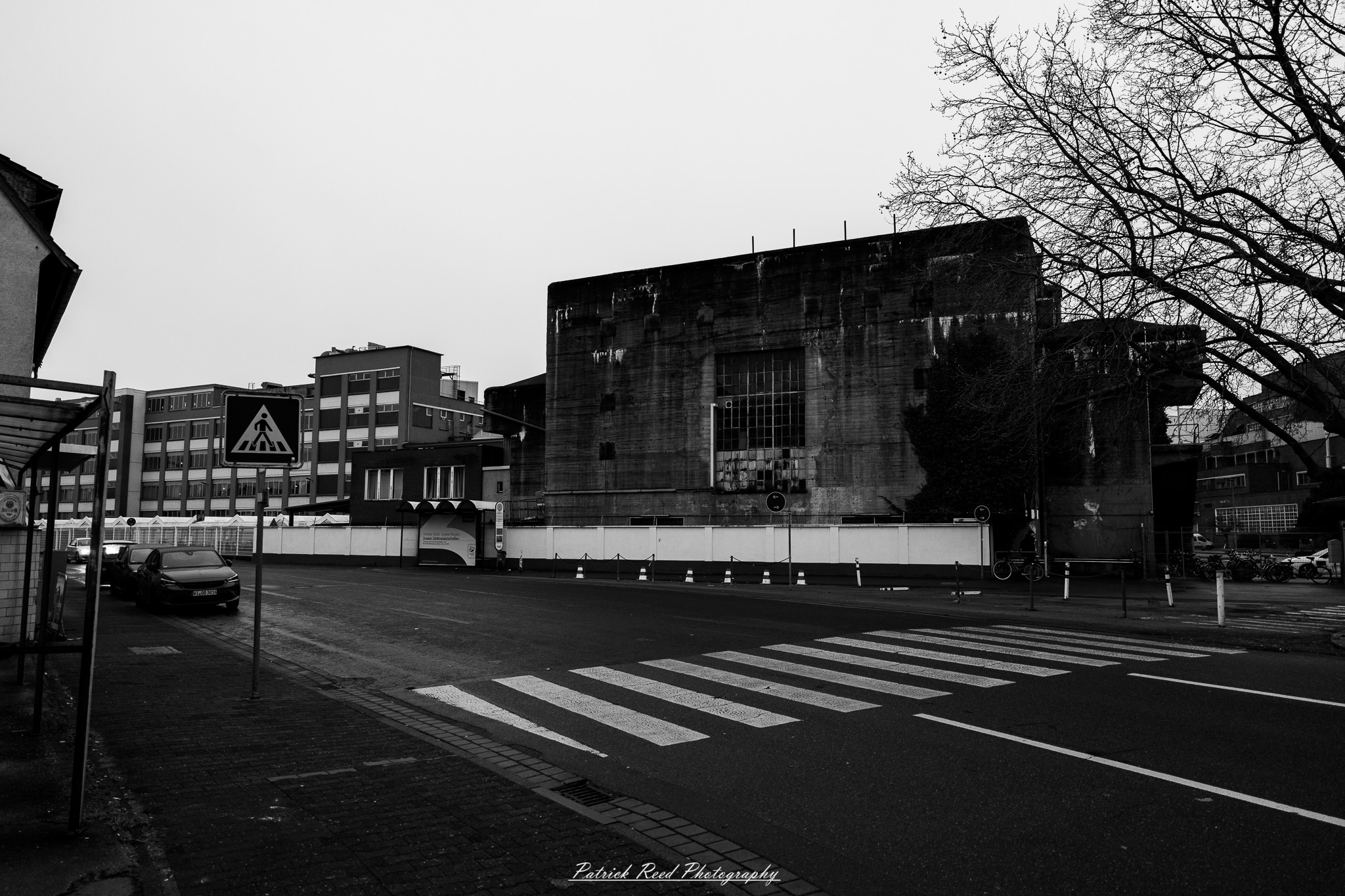 Russelsheim 4 A series of noir-style street photography scenes set in Rüsselsheim, Germany, at night. The dimly lit streets feature wet cobblestones reflecting the glow of streetlights and neon signs. Shadows and silhouettes of people, some walking or riding bicycles, add a mysterious atmosphere. Industrial architecture, such as factory buildings and modern urban structures, contrasts with old-world charm. Elements like parked cars, lampposts, and occasional graffiti create a gritty, cinematic ambiance. The misty air and moody lighting emphasize the noir aesthetic, evoking a sense of intrigue and storytelling.