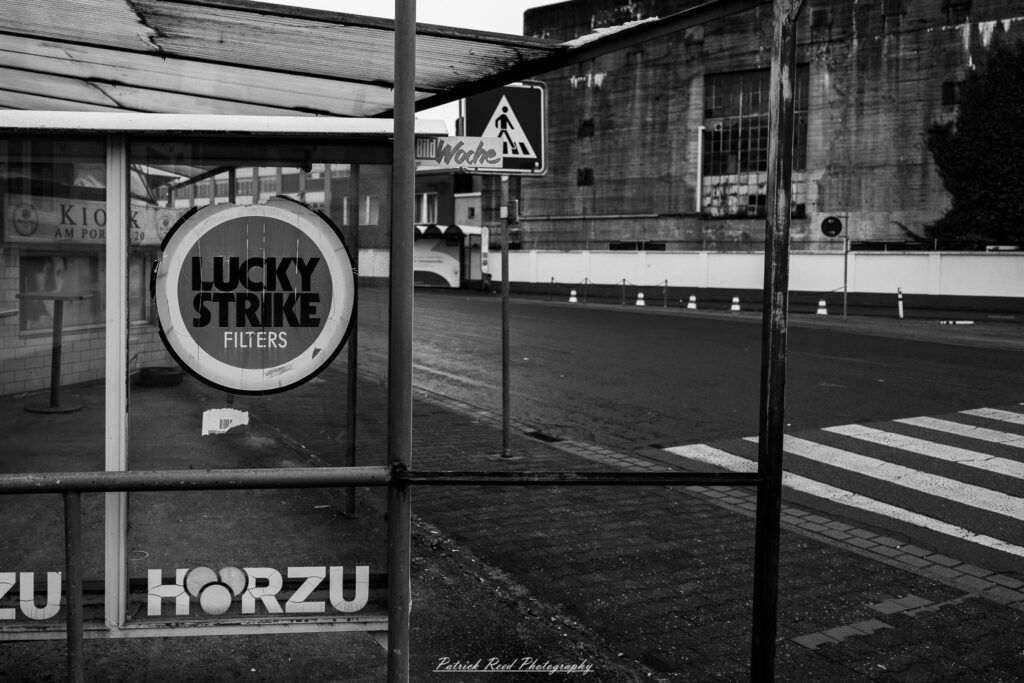 A series of noir-style street photography scenes set in Rüsselsheim, Germany, at night. The dimly lit streets feature wet cobblestones reflecting the glow of streetlights and neon signs. Shadows and silhouettes of people, some walking or riding bicycles, add a mysterious atmosphere. Industrial architecture, such as factory buildings and modern urban structures, contrasts with old-world charm. Elements like parked cars, lampposts, and occasional graffiti create a gritty, cinematic ambiance. The misty air and moody lighting emphasize the noir aesthetic, evoking a sense of intrigue and storytelling.