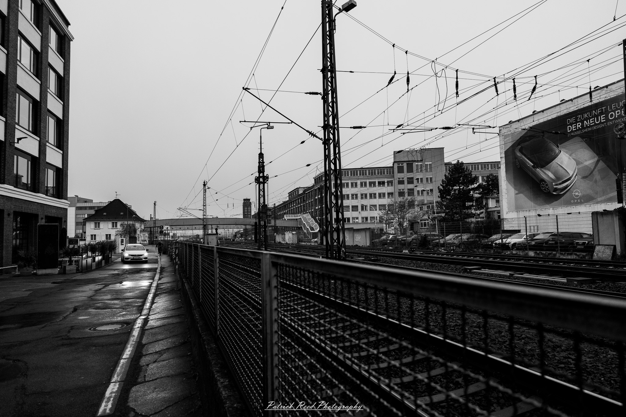 Russelsheim 7 A series of noir-style street photography scenes set in Rüsselsheim, Germany, at night. The dimly lit streets feature wet cobblestones reflecting the glow of streetlights and neon signs. Shadows and silhouettes of people, some walking or riding bicycles, add a mysterious atmosphere. Industrial architecture, such as factory buildings and modern urban structures, contrasts with old-world charm. Elements like parked cars, lampposts, and occasional graffiti create a gritty, cinematic ambiance. The misty air and moody lighting emphasize the noir aesthetic, evoking a sense of intrigue and storytelling.