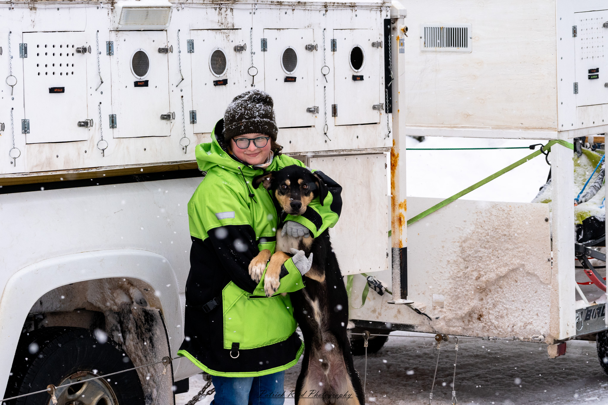 A team of sled dogs charges through a snow-covered landscape, their harnesses taut as they pull the sled forward. The musher, bundled in thick winter gear, stands at the back, steering through the icy terrain. Their breath mingles with the cold air, and snowflakes swirl around them. Towering, frost-covered trees line the trail, with distant mountains barely visible through the wintry haze. The muted winter light casts long shadows, adding to the sense of adventure and resilience in this frozen wilderness.