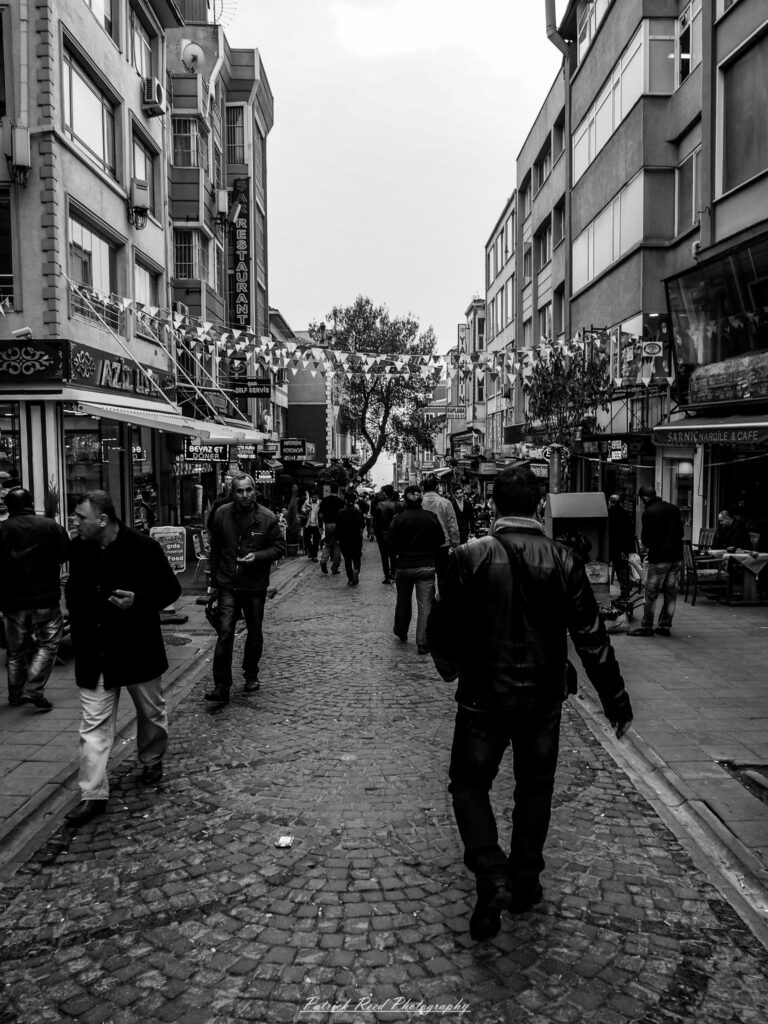 A series of noir-style street photography scenes set in Istanbul, Turkey, at night. The dimly lit streets and narrow alleys are wet from rain, reflecting the warm glow of streetlights and neon signs. Silhouettes of people walking, some carrying umbrellas, move through the misty atmosphere. The scenes highlight a blend of historical Ottoman architecture, such as arched doorways and intricate stone facades, with modern urban elements like cafes, storefronts, and graffiti. The Bosphorus skyline and glimpses of minarets in the distance add to the cinematic, mysterious ambiance. The overall mood is evocative and steeped in noir storytelling.