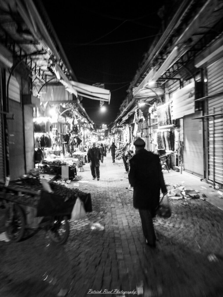A series of noir-style street photography scenes set in Istanbul, Turkey, at night. The dimly lit streets and narrow alleys are wet from rain, reflecting the warm glow of streetlights and neon signs. Silhouettes of people walking, some carrying umbrellas, move through the misty atmosphere. The scenes highlight a blend of historical Ottoman architecture, such as arched doorways and intricate stone facades, with modern urban elements like cafes, storefronts, and graffiti. The Bosphorus skyline and glimpses of minarets in the distance add to the cinematic, mysterious ambiance. The overall mood is evocative and steeped in noir storytelling.