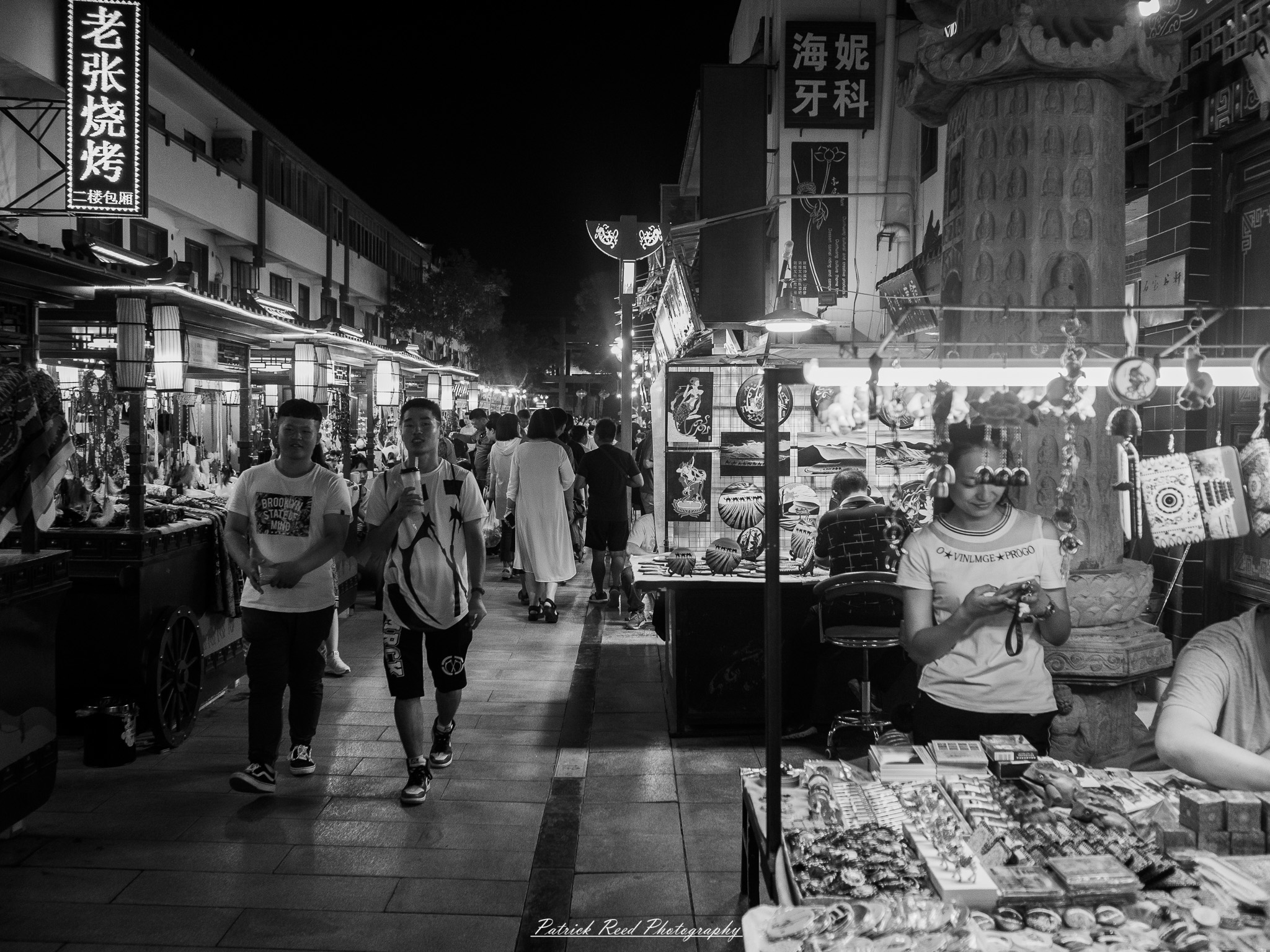 A series of noir-style street photography scenes set in Dunhuang, China, at night. The dimly lit streets and narrow alleyways reflect the soft glow of lanterns and neon signs from small shops and market stalls. Silhouettes of people walk through the misty night, some in traditional robes, adding a sense of mystery. The historic Silk Road atmosphere is highlighted by earthen architecture, intricate wooden doors, and desert dust settling on the pavement. Modern elements like motorbikes and distant headlights contrast with the ancient setting. The deep shadows and warm, flickering lights create a cinematic, moody ambiance, blending noir aesthetics with Dunhuang’s rich cultural history.